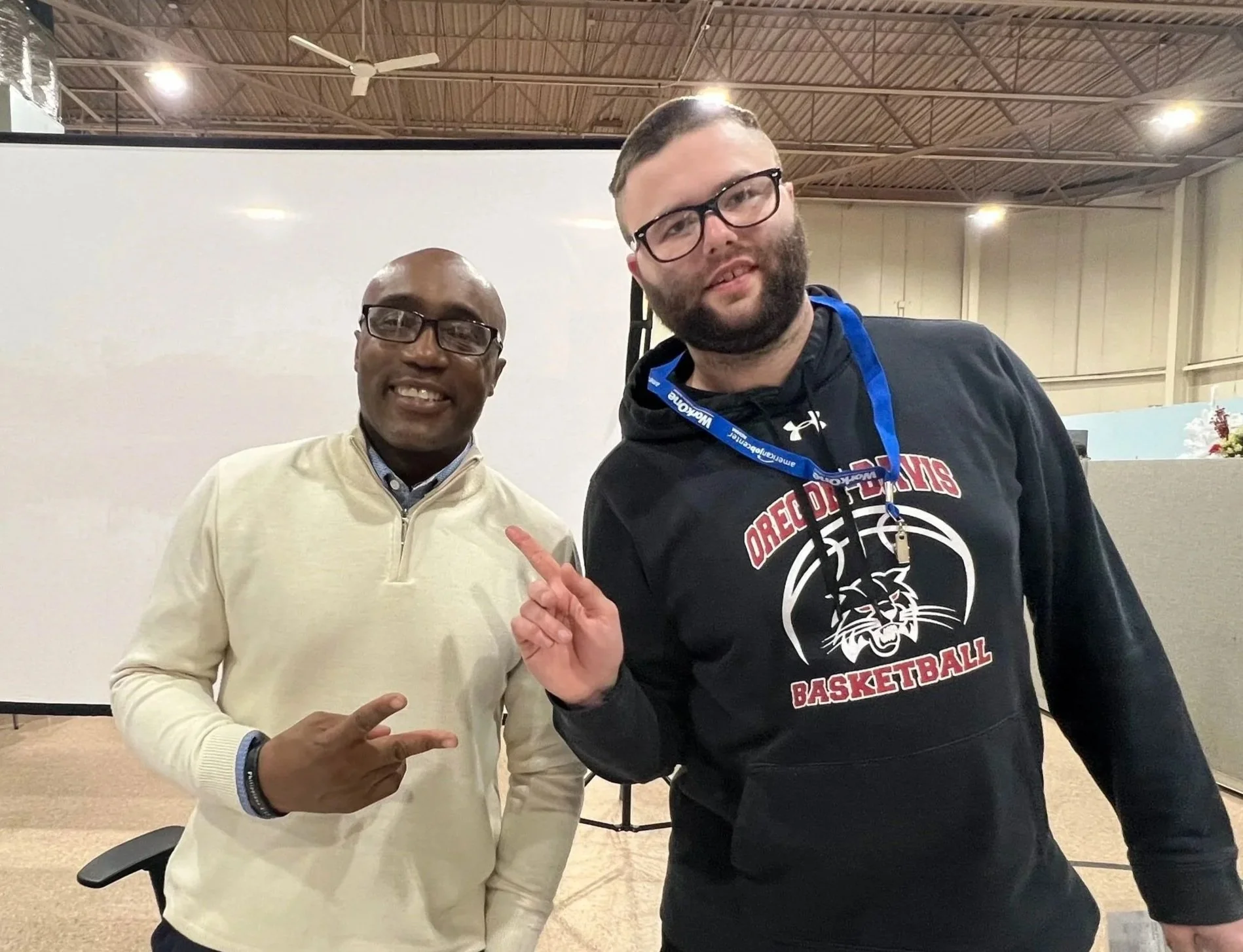 Two men standing inside a building, smiling at the camera. The man on the left is wearing glasses and a cream-colored sweater, pointing towards the man on the right. The man on the right is wearing glasses, a hoodie with a basketball logo, and an honor cord, with a lanyard around his neck. The background includes a blank wall and a ceiling with a fan.