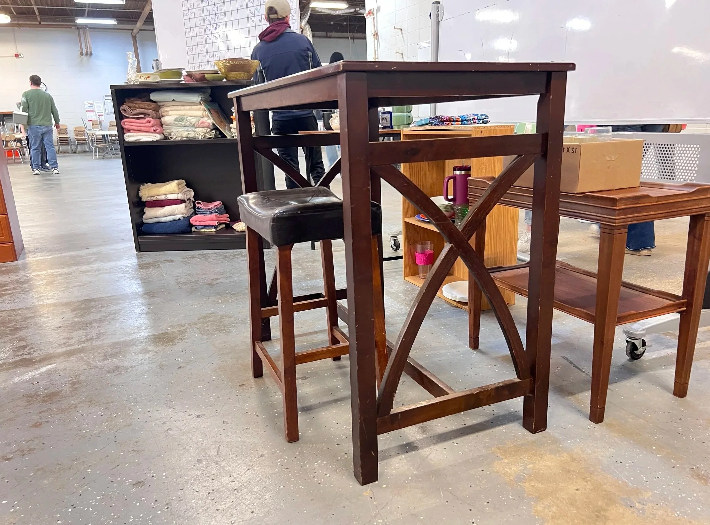 Indoor setting with furniture including a tall wooden table with a black cushioned stool, a black shelf with neatly folded towels, and a small brown tray table. People are visible in the background, one wearing a hat and jacket and others walking.