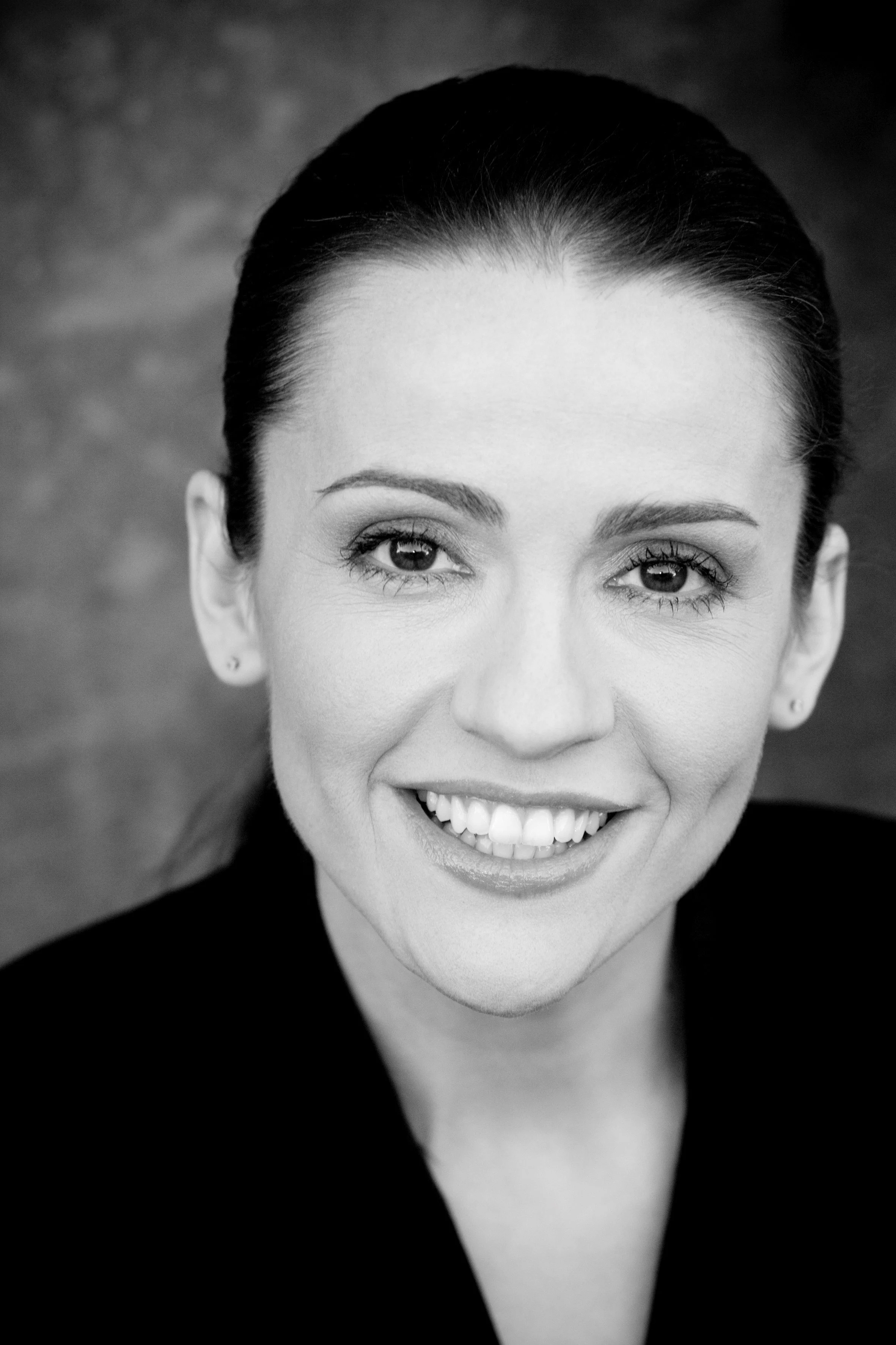 Black and white portrait of a smiling woman with dark hair pulled back, light skin, and bright eyes, wearing a dark top.