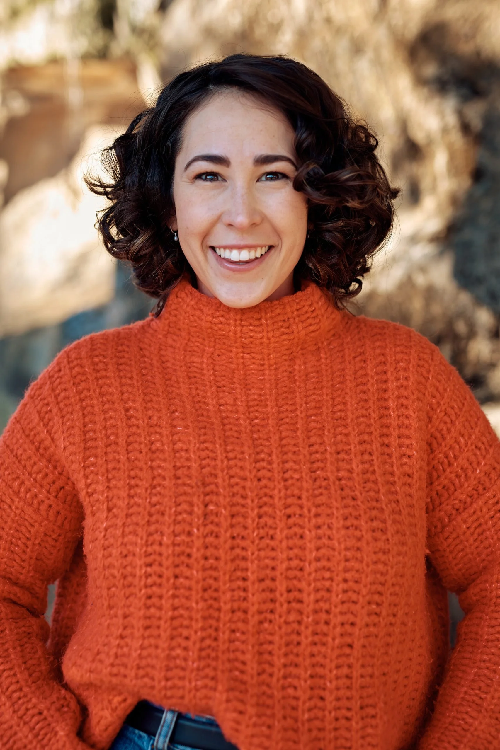 A woman smiling outdoors in front of a blurred background, wearing an orange knit sweater.