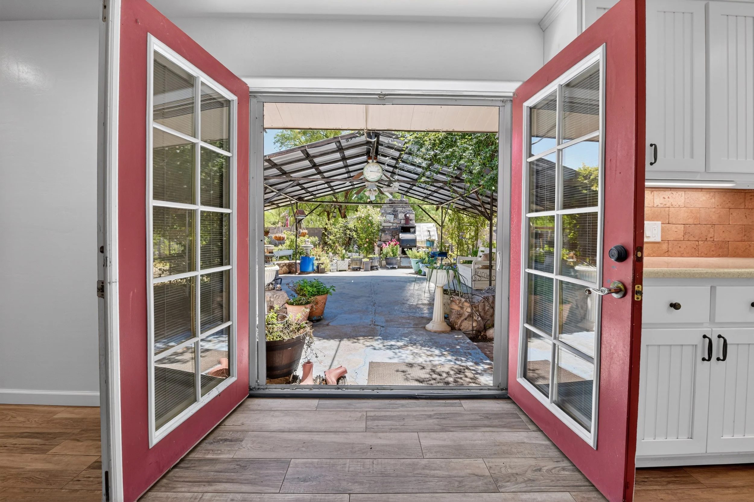 View of an outdoor patio through open red double doors with glass panes, showing a roofed area with a clock, plants, garden tools, and outdoor furniture.