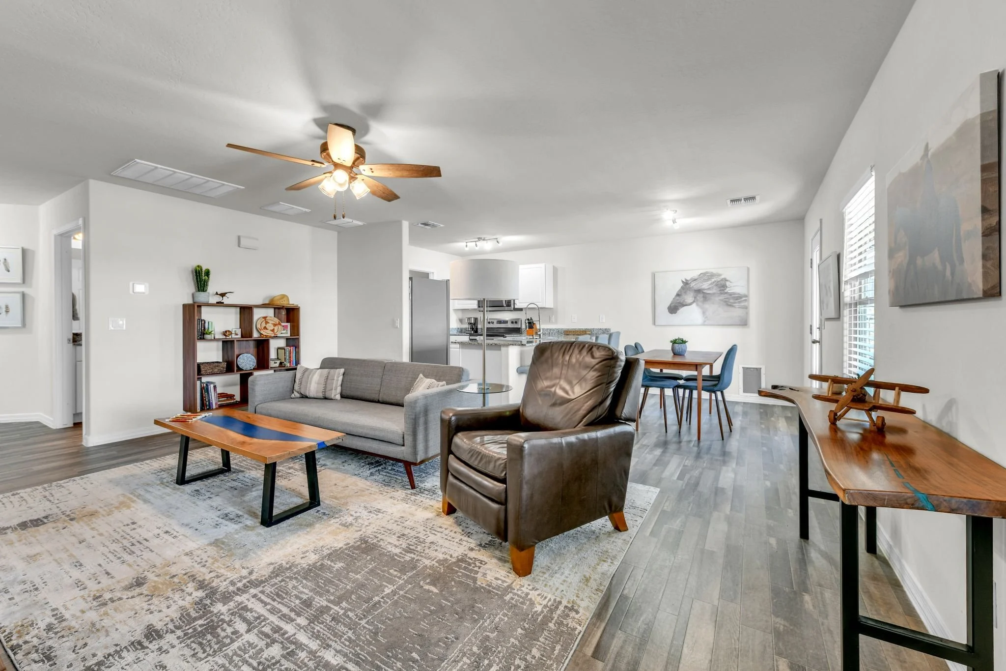 A spacious living room with a white ceiling fan, light-colored walls, and hardwood flooring. The room features a gray sofa, a leather armchair, a wooden coffee table, a rug, a bookshelf with decorative items, and a dining area with a wooden table and