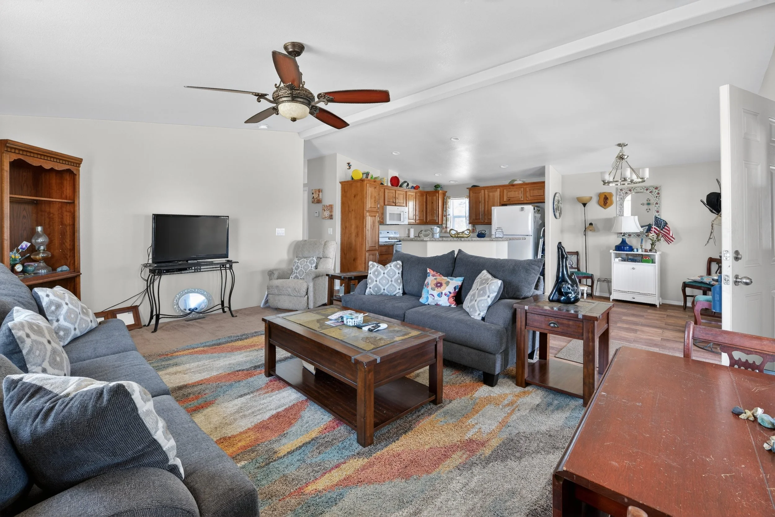A living room with gray sofas, a wooden coffee table, a colorful area rug, and a ceiling fan. The background shows a kitchen with wooden cabinets and white appliances.