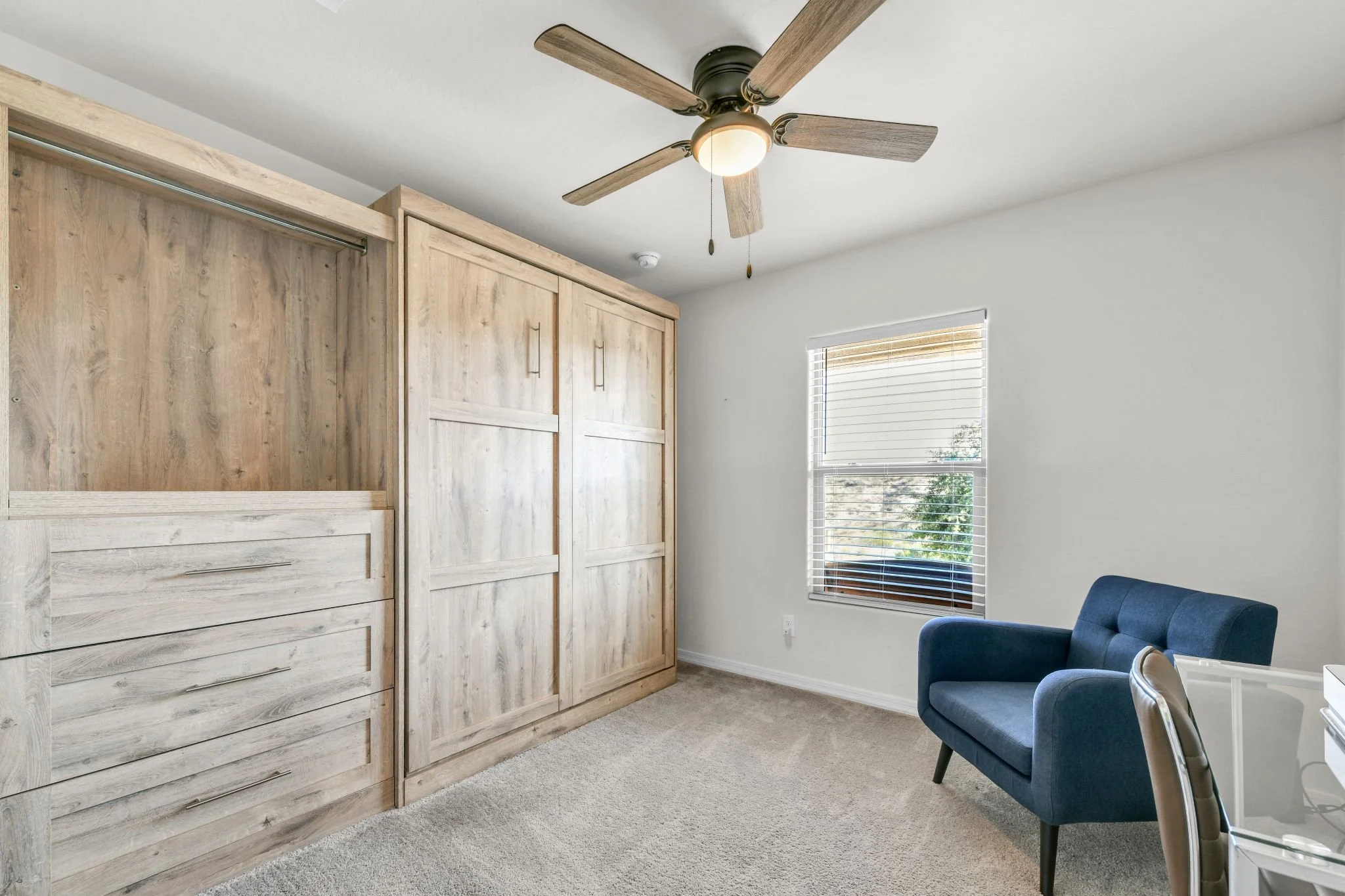 Interior of a room with a wooden wardrobe, a ceiling fan with wooden blades, a window with blinds, and a blue armchair.
