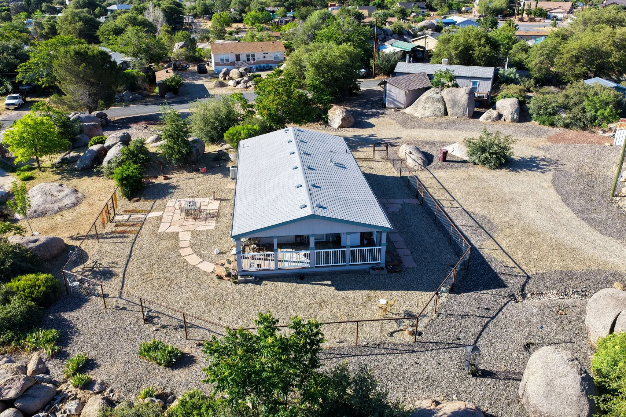An aerial view of a residential backyard with a white metal roof building, surrounded by a gravel yard, trees, bushes, large rocks, and a fenced area with outdoor furniture.