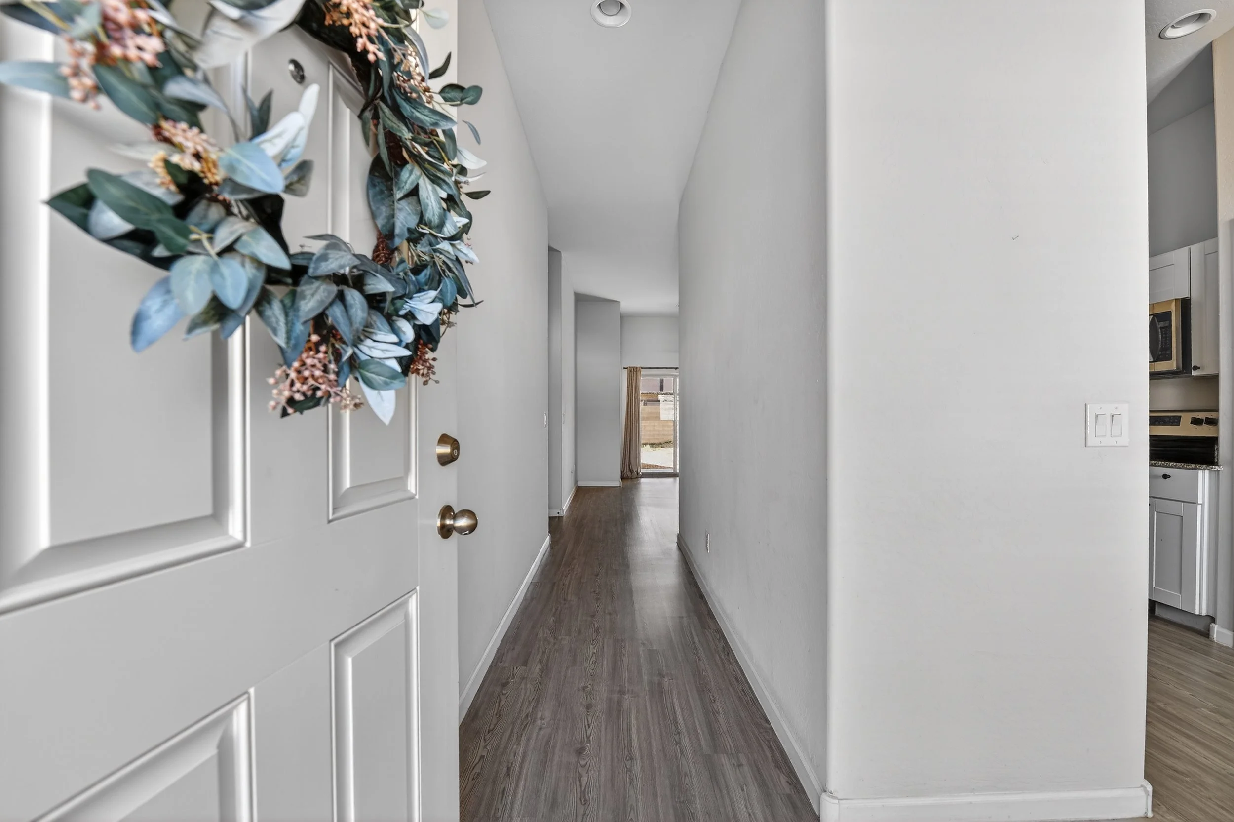 Inside view of a hallway with white walls and wooden flooring, decorated with a floral wreath on the door on the left side, leading to a room with window and curtains.