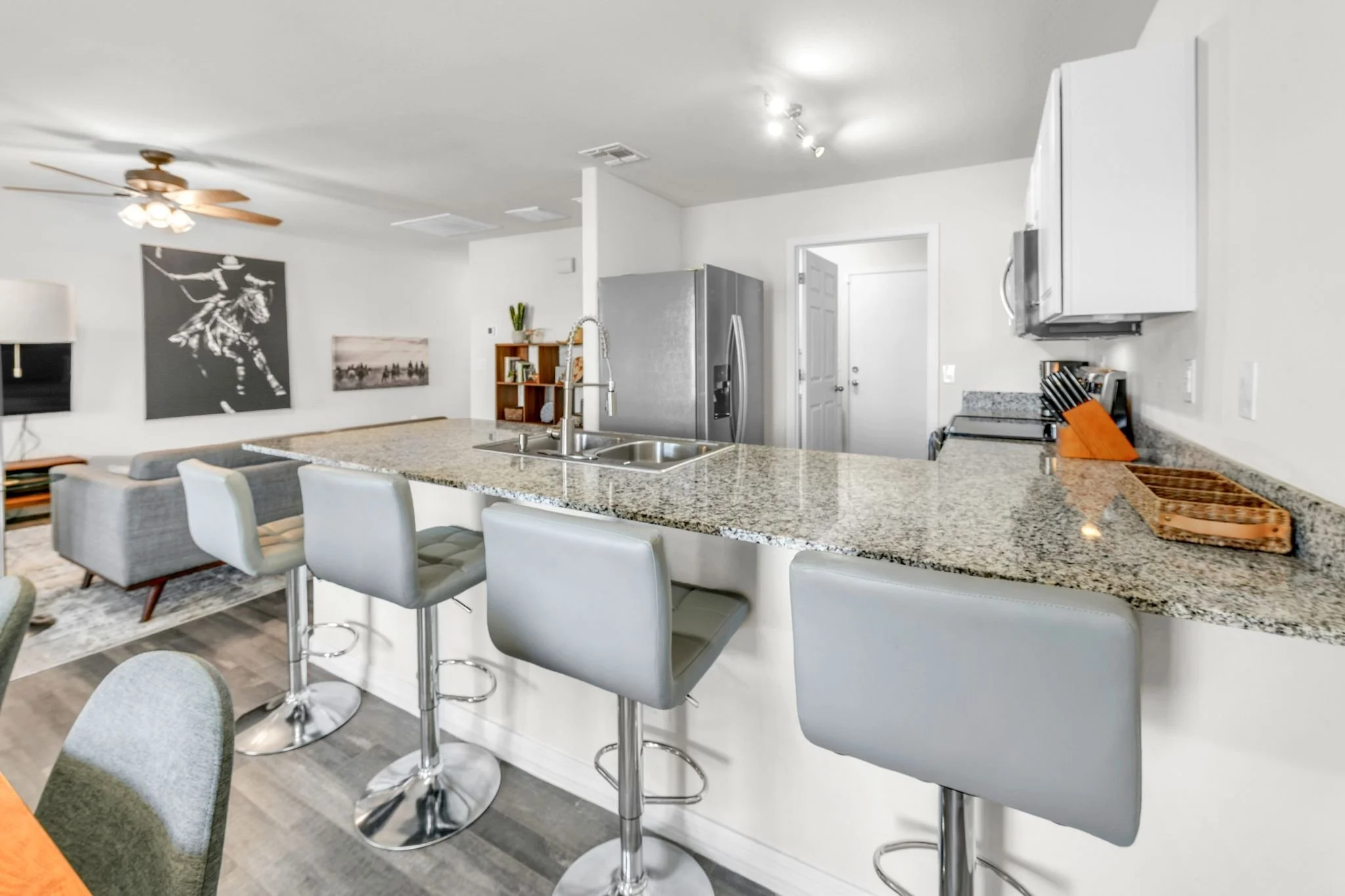 A modern kitchen with a granite island, white cabinets, a stainless steel refrigerator, and barstools. The background features wall art and a ceiling fan.