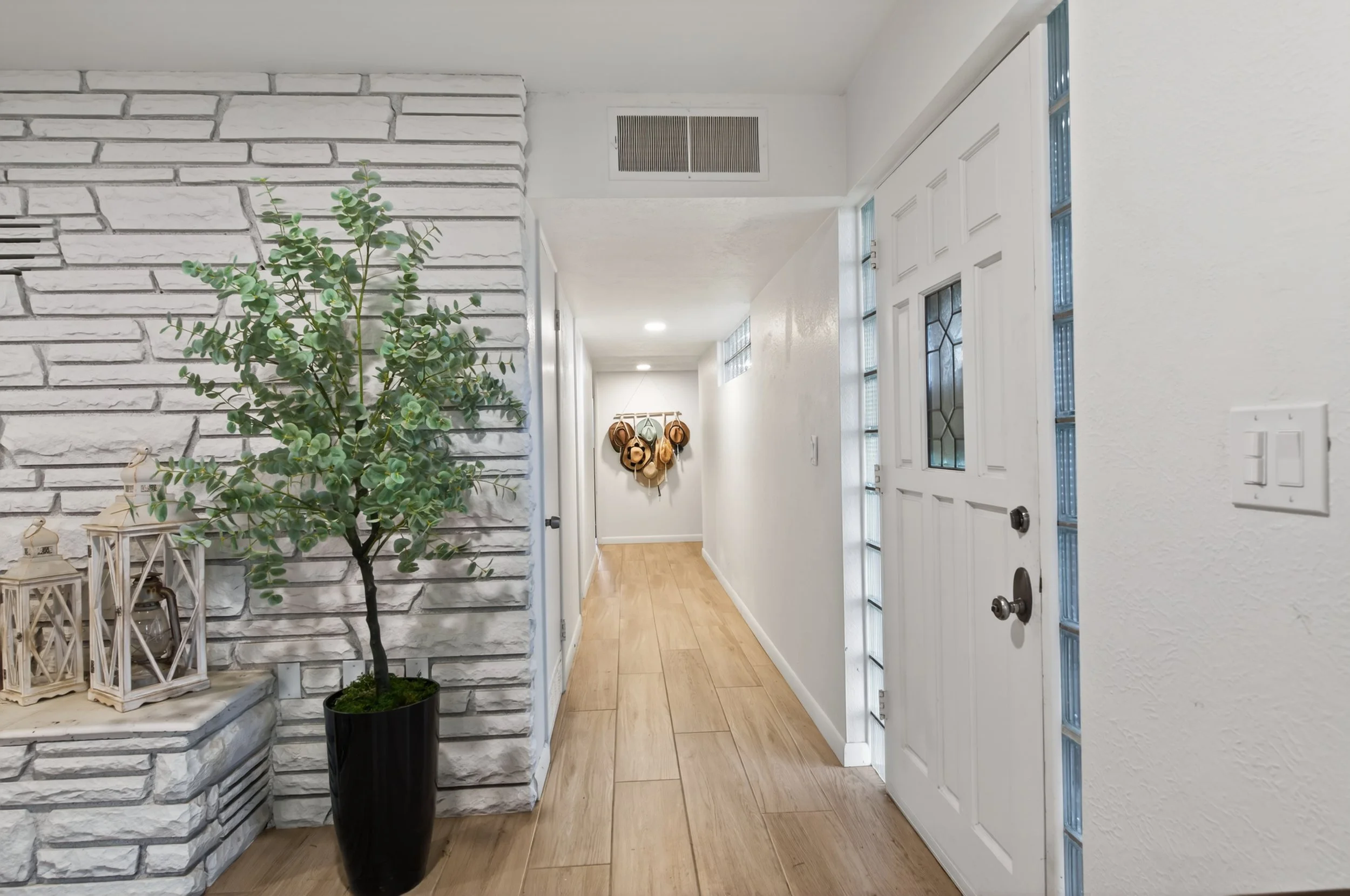 Interior view of a hallway with white textured walls, a white front door with glass panels, a potted plant, and decorative lanterns near a white brick accent wall.