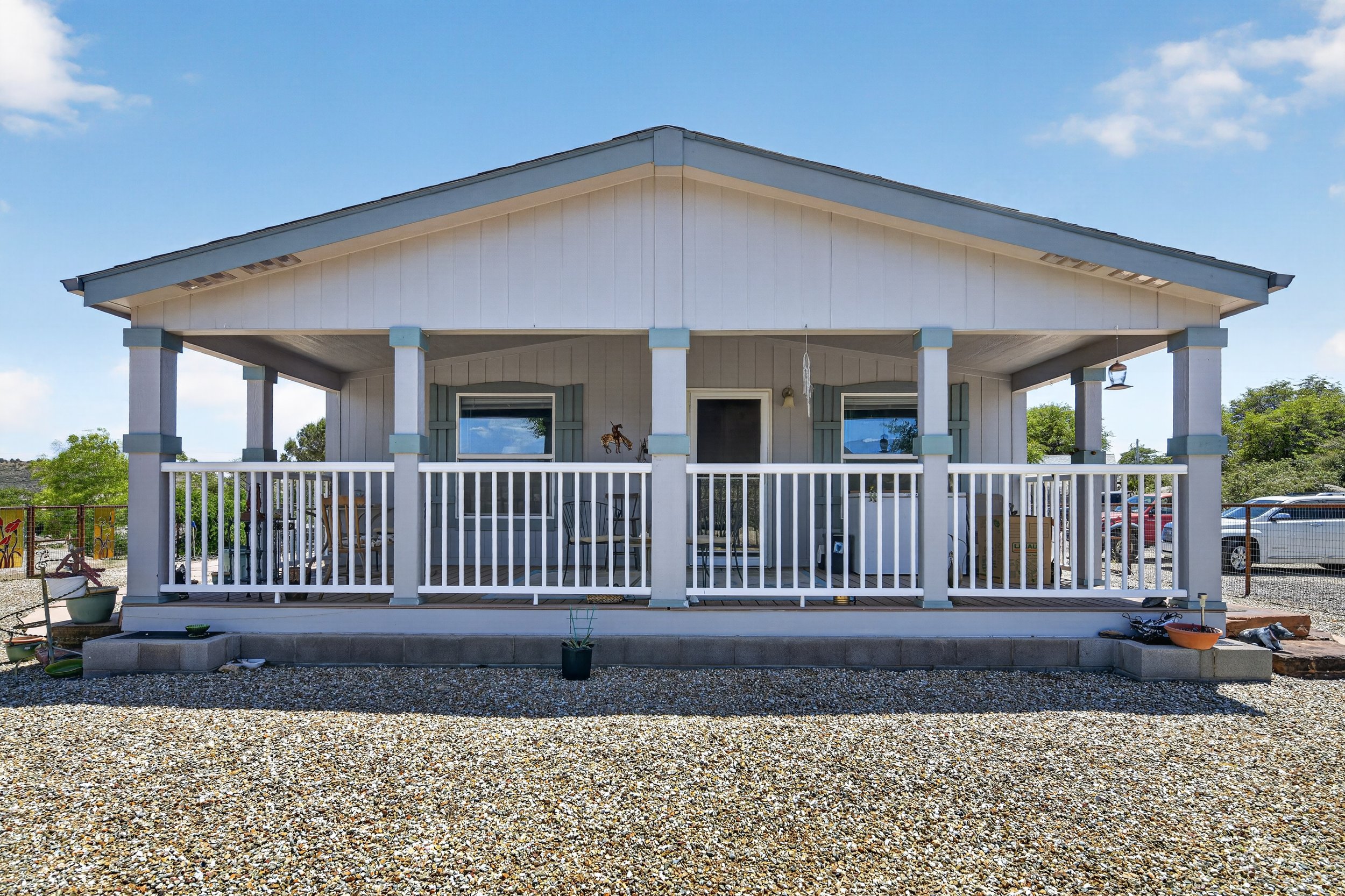 Front view of a white house with a porch, small hanging light, and two windows with shutters, surrounded by a gravel yard and blue sky.