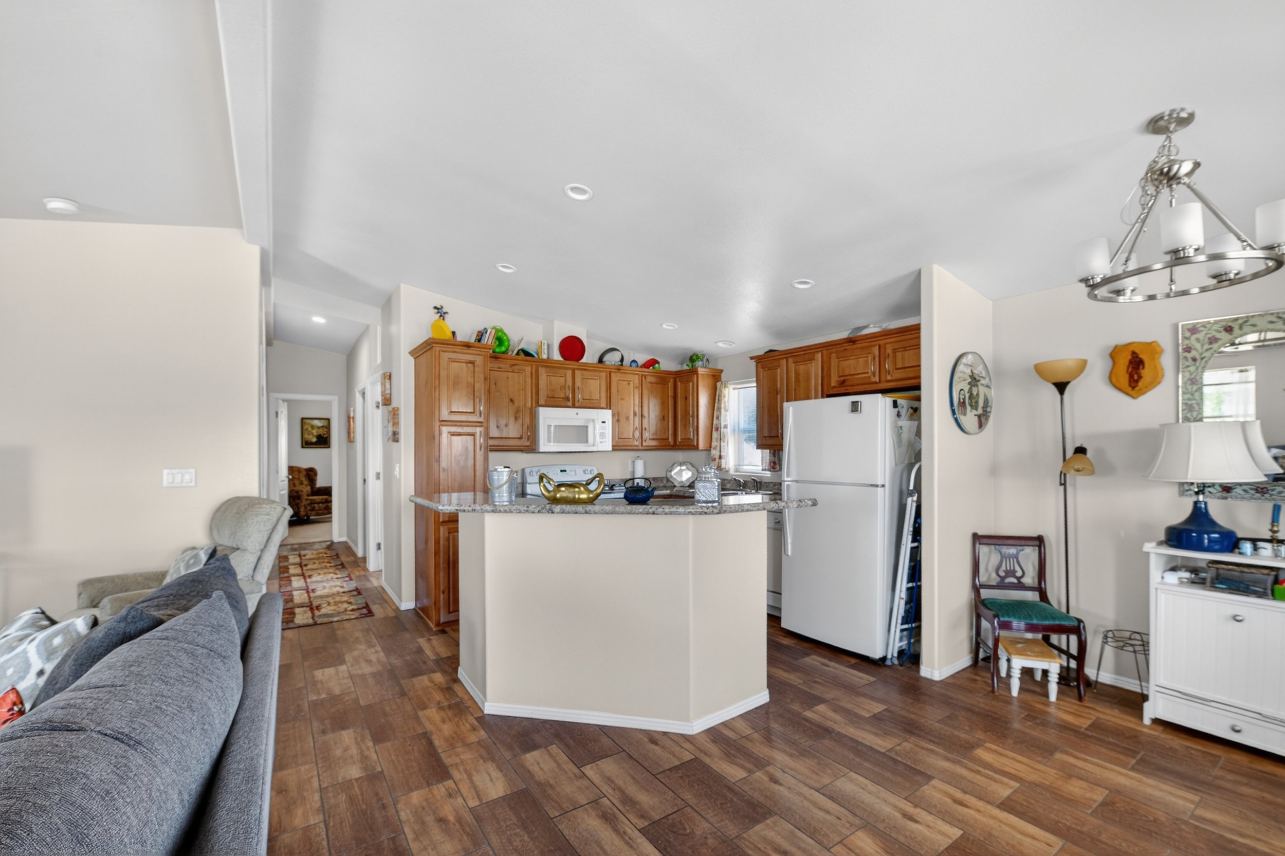 Open-concept kitchen and living room with wooden cabinets, white refrigerator, and a granite island. The living area has a gray couch, a patterned rug, and a decorative lamp, with wood flooring throughout.