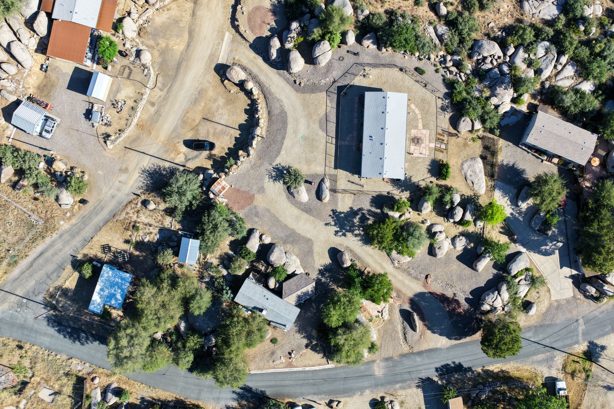 An aerial view of a residential area with houses, trees, and large rocks. There are paved roads and driveways, along with some vehicles parked near the houses.