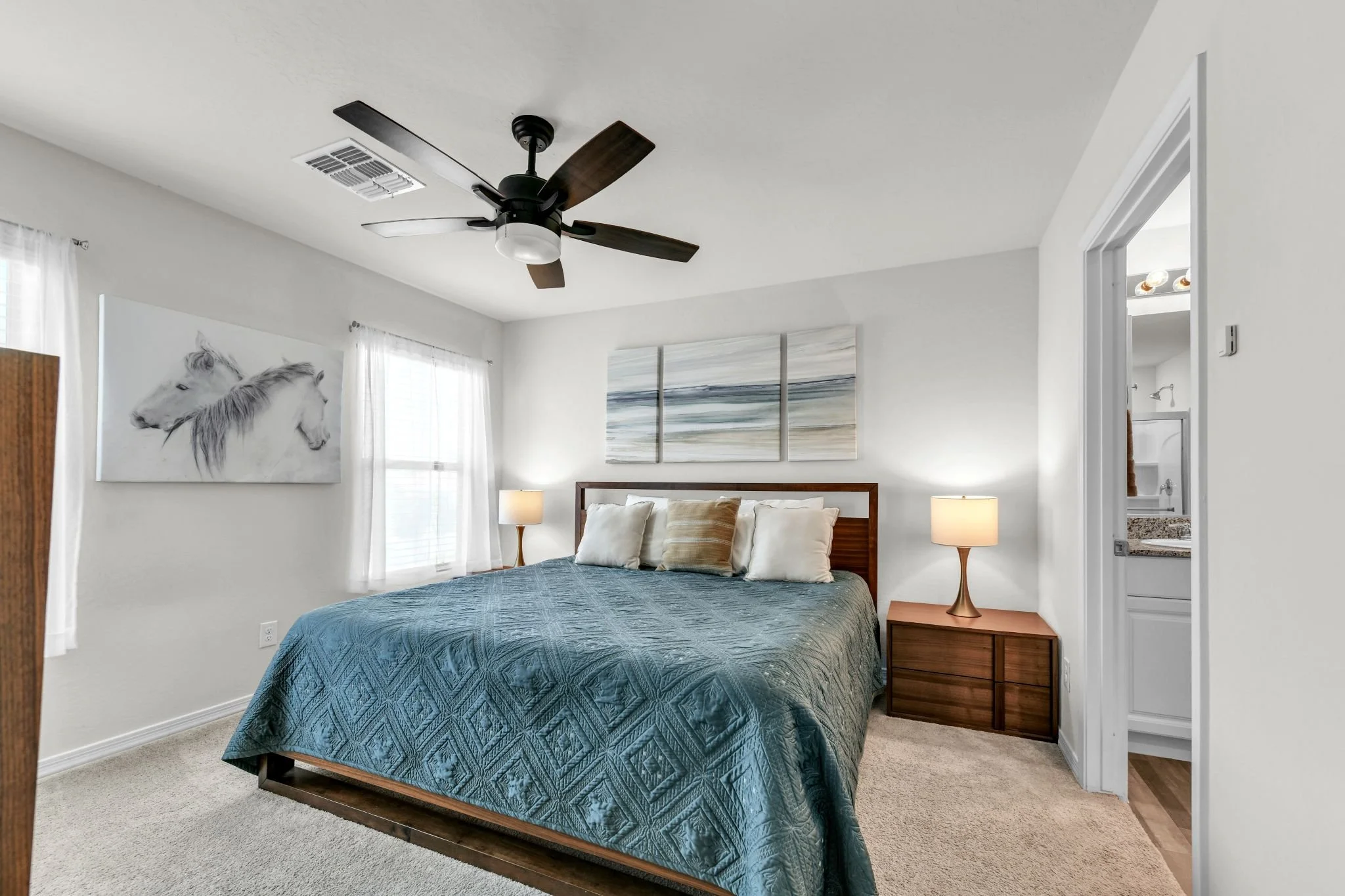 Bright bedroom with a wooden bed frame, teal quilt, and pillows. Artwork of a white horse hangs above the bed. Two lamps sit on wooden nightstands. A ceiling fan with dark blades is on the ceiling. White curtains on windows let in natural light. An o