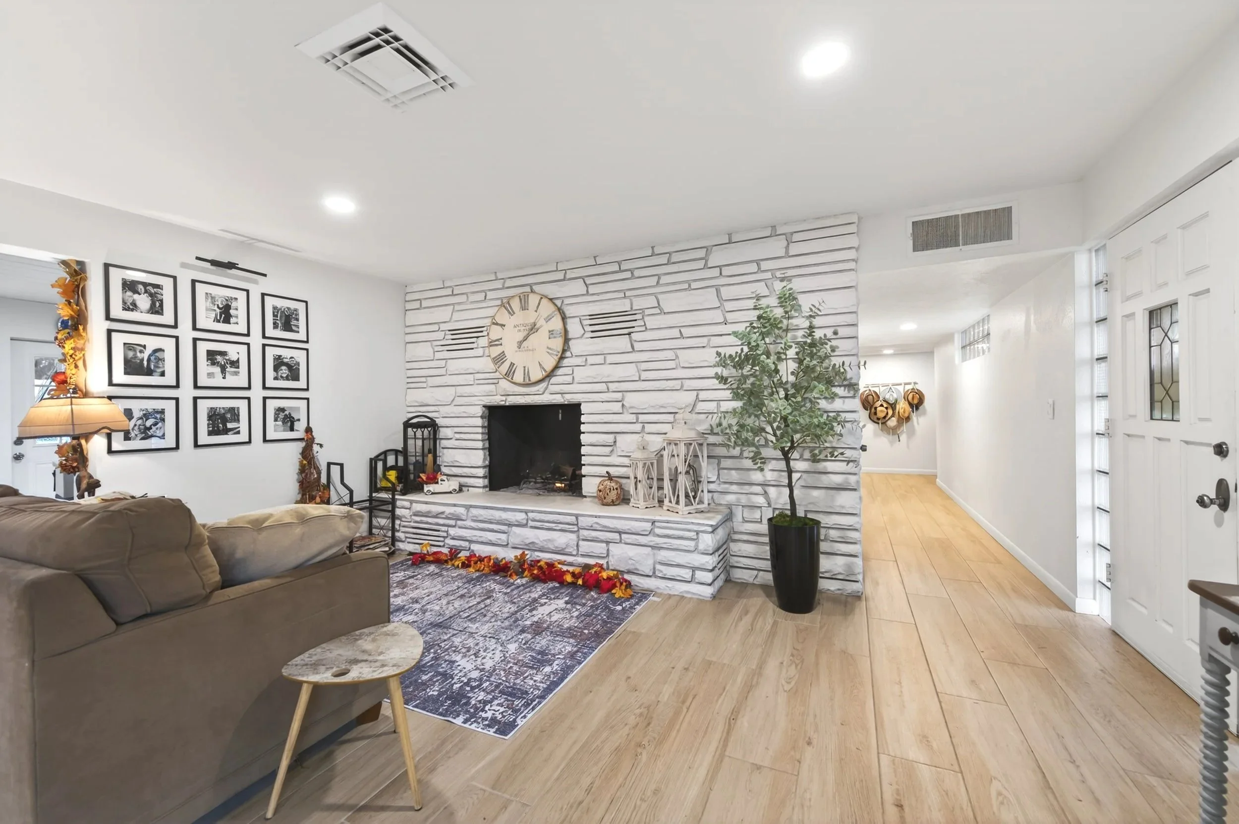 Living room with white brick fireplace, large wall clock, beige couch, black framed photo gallery, potted plant, decorative lanterns and fall leaf arrangement.