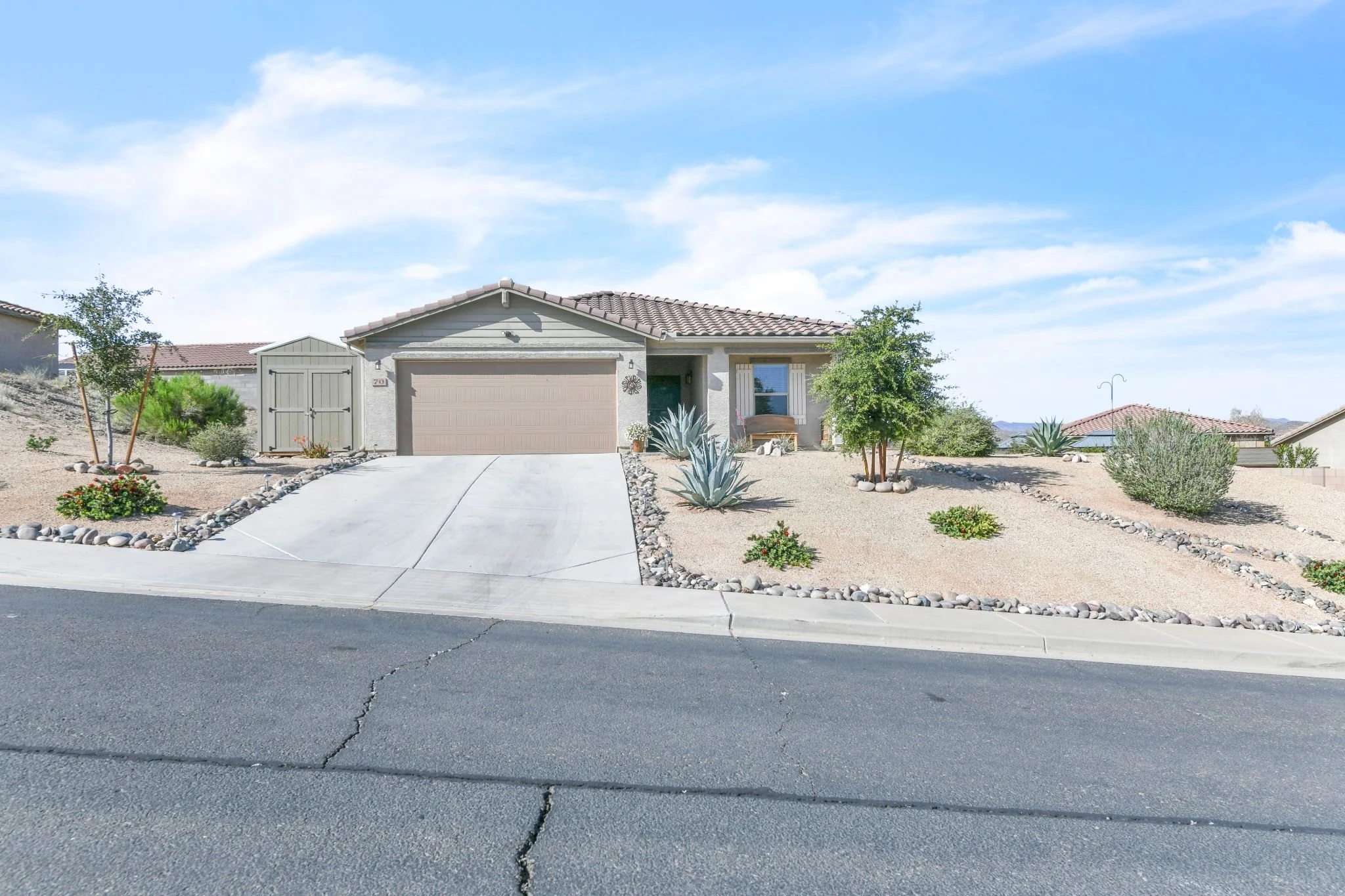 A suburban house with a beige garage door, decorative plants, and a desert landscape yard with rocks and drought-tolerant plants under a blue sky.