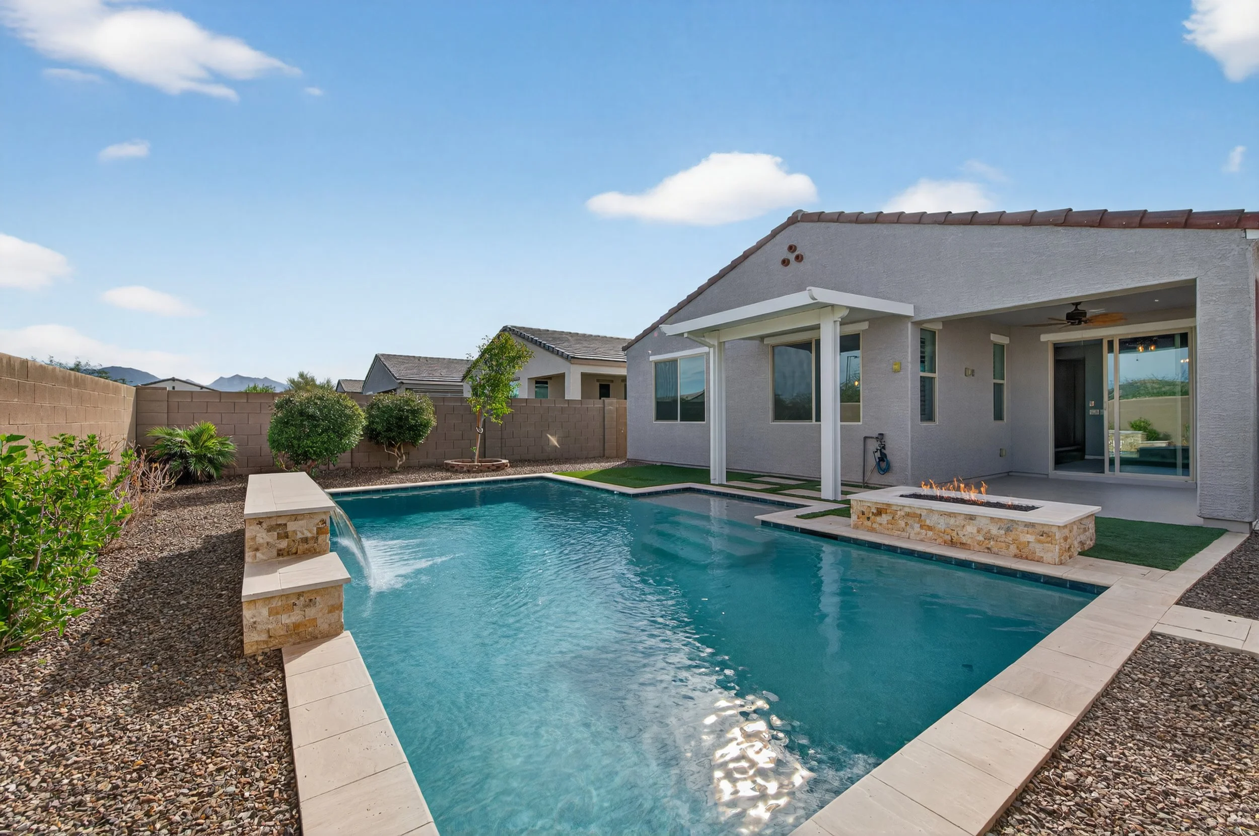 Backyard with a swimming pool, some trees, and a house with a patio under a blue sky with clouds.