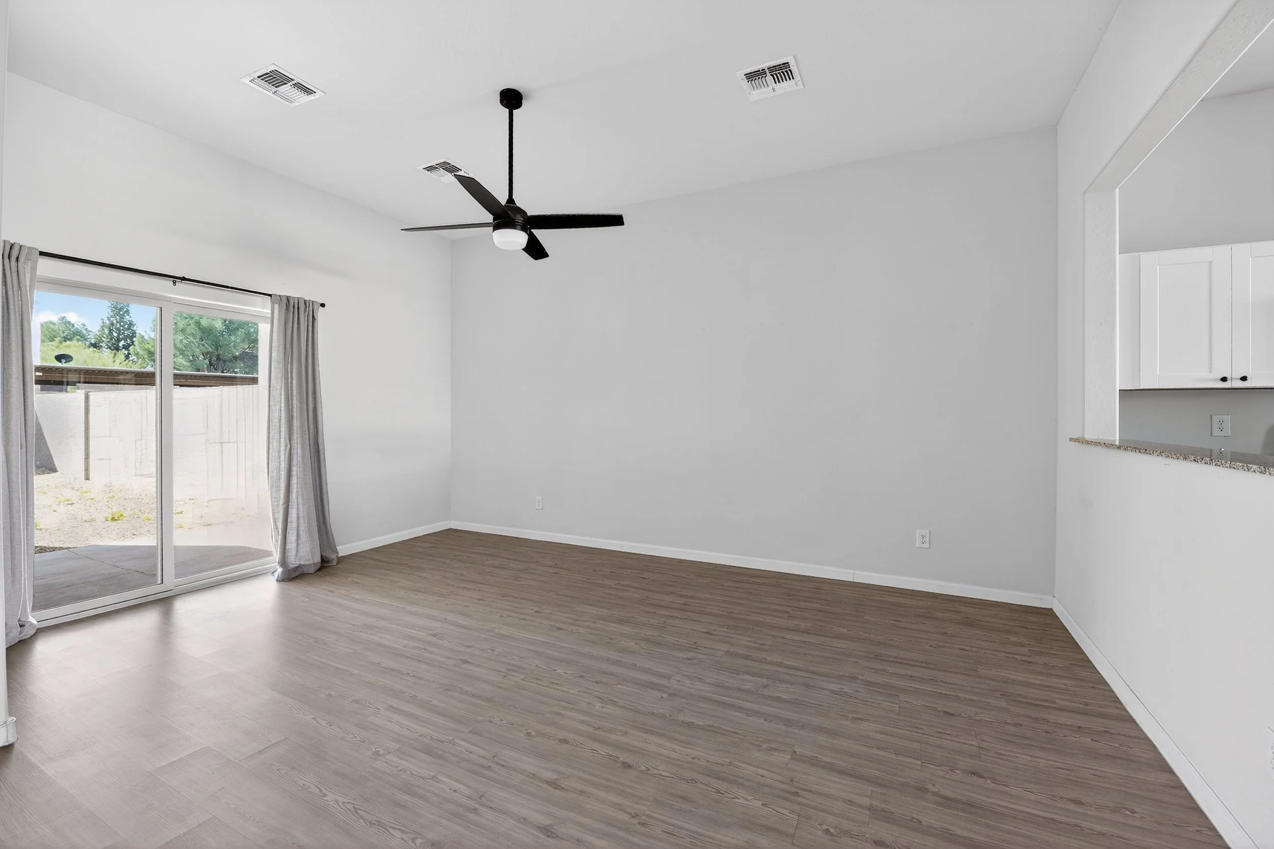 Empty living room with light-colored wood flooring, white walls, a sliding glass door with curtains, a black ceiling fan with a light, and part of a white kitchen cabinet visible.