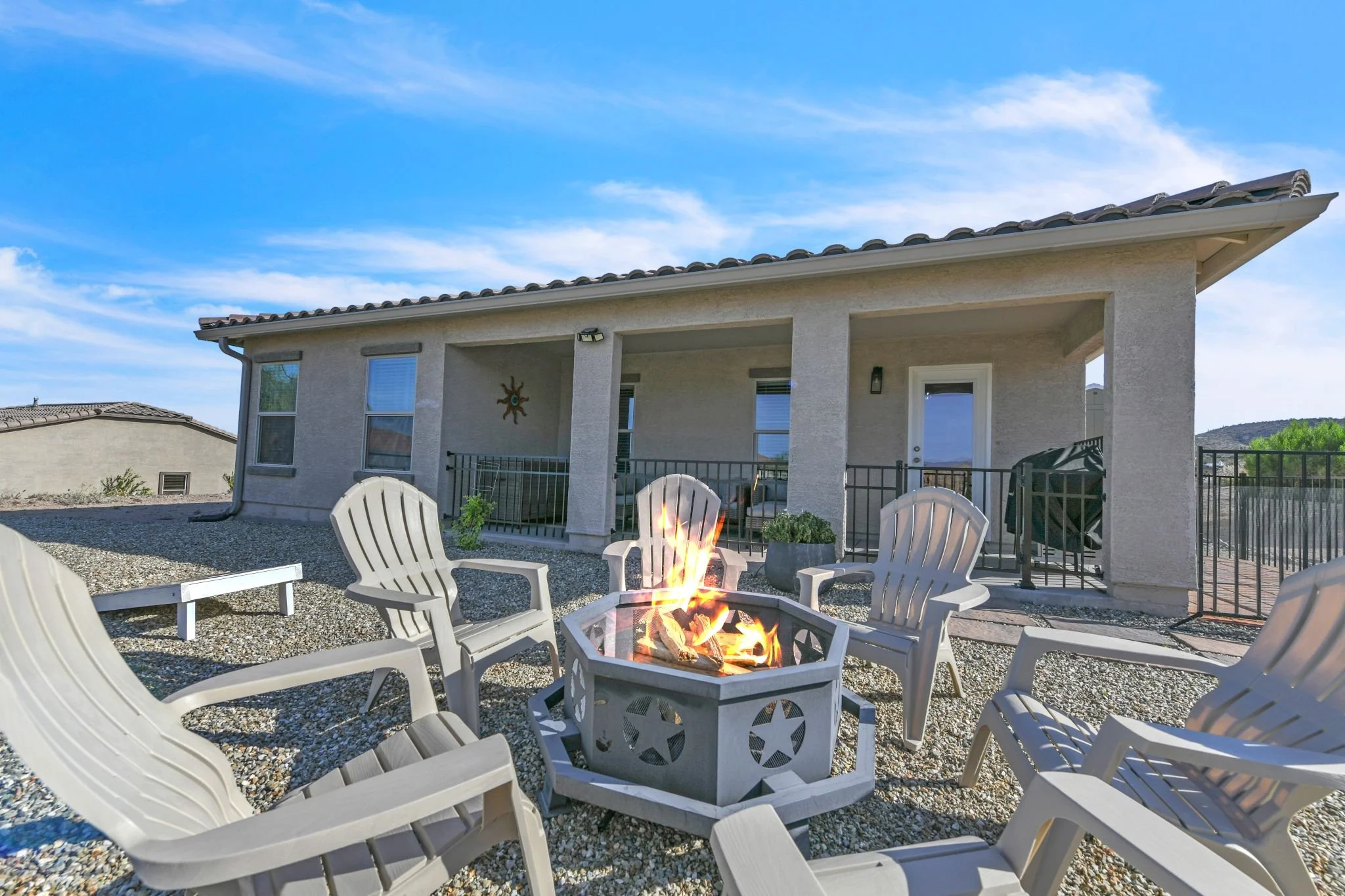 Backyard patio area with a firepit surrounded by white chairs, a covered porch with a wall decoration, and neighboring houses under a blue sky with clouds.