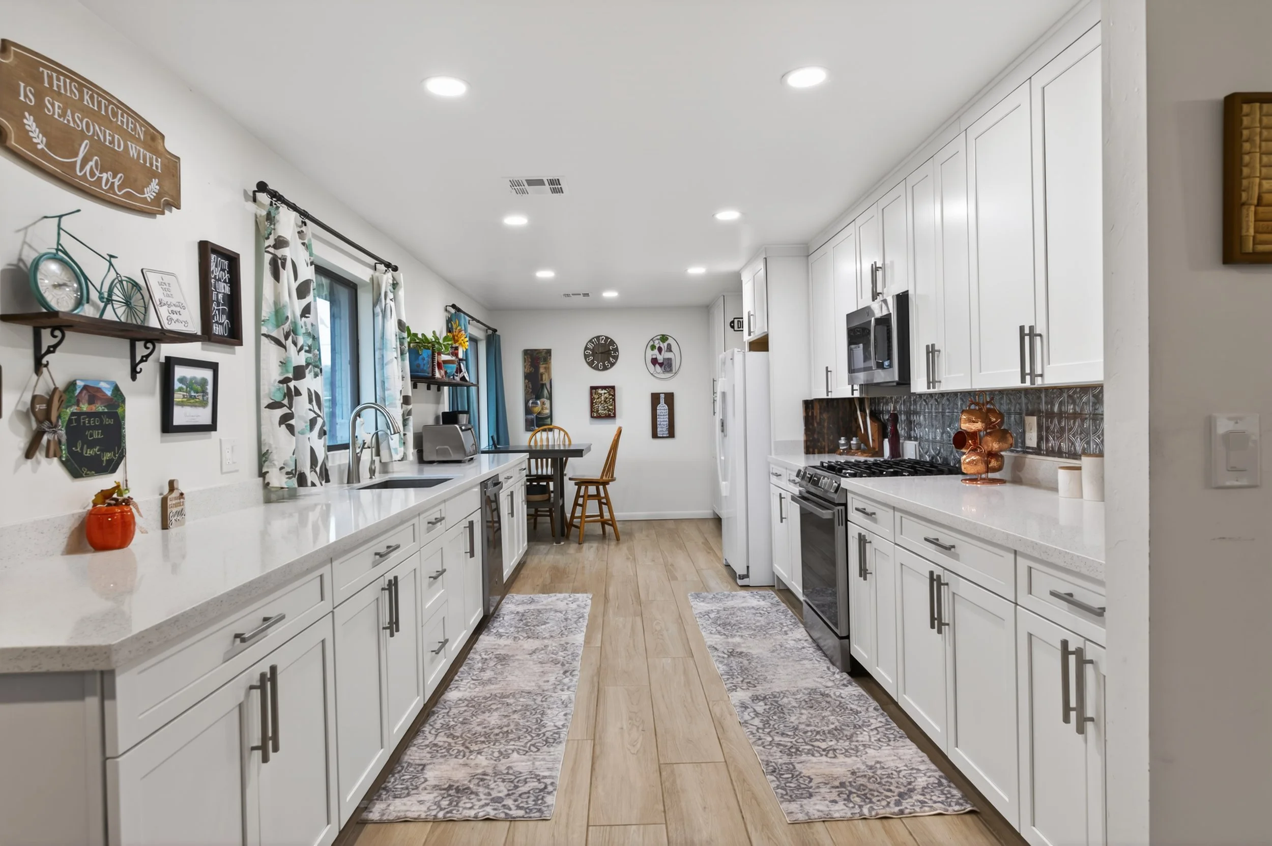 Bright modern kitchen with white cabinets, countertops, stainless steel appliances, and decorative items on the walls and shelves, featuring a window with patterned curtains and wooden flooring.