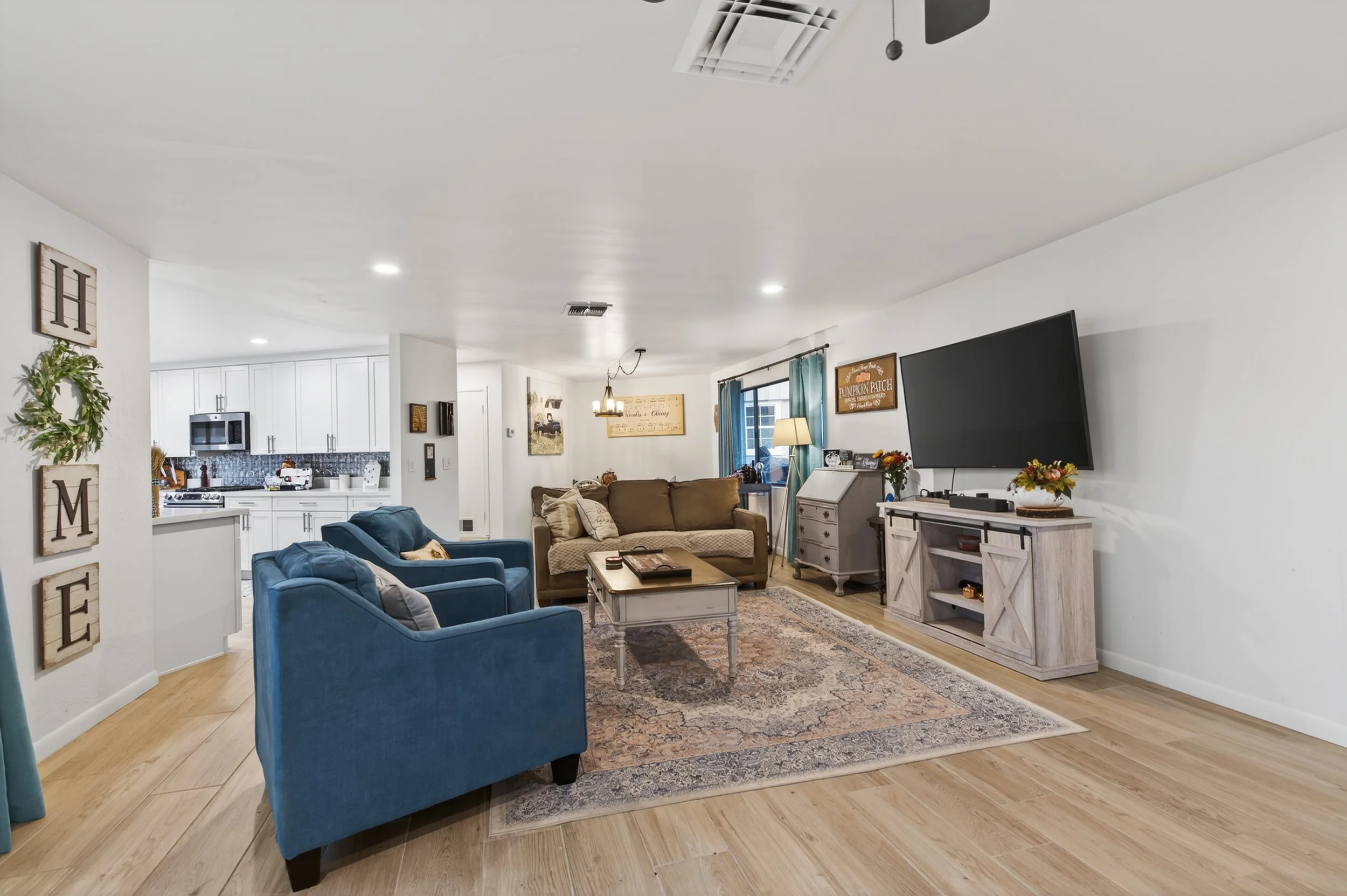 A spacious living room with hardwood floors, featuring a blue armchair and a brown sofa arranged around a coffee table. The room includes white walls, a large flat-screen TV mounted on the wall, and various decorative elements, including framed artwo