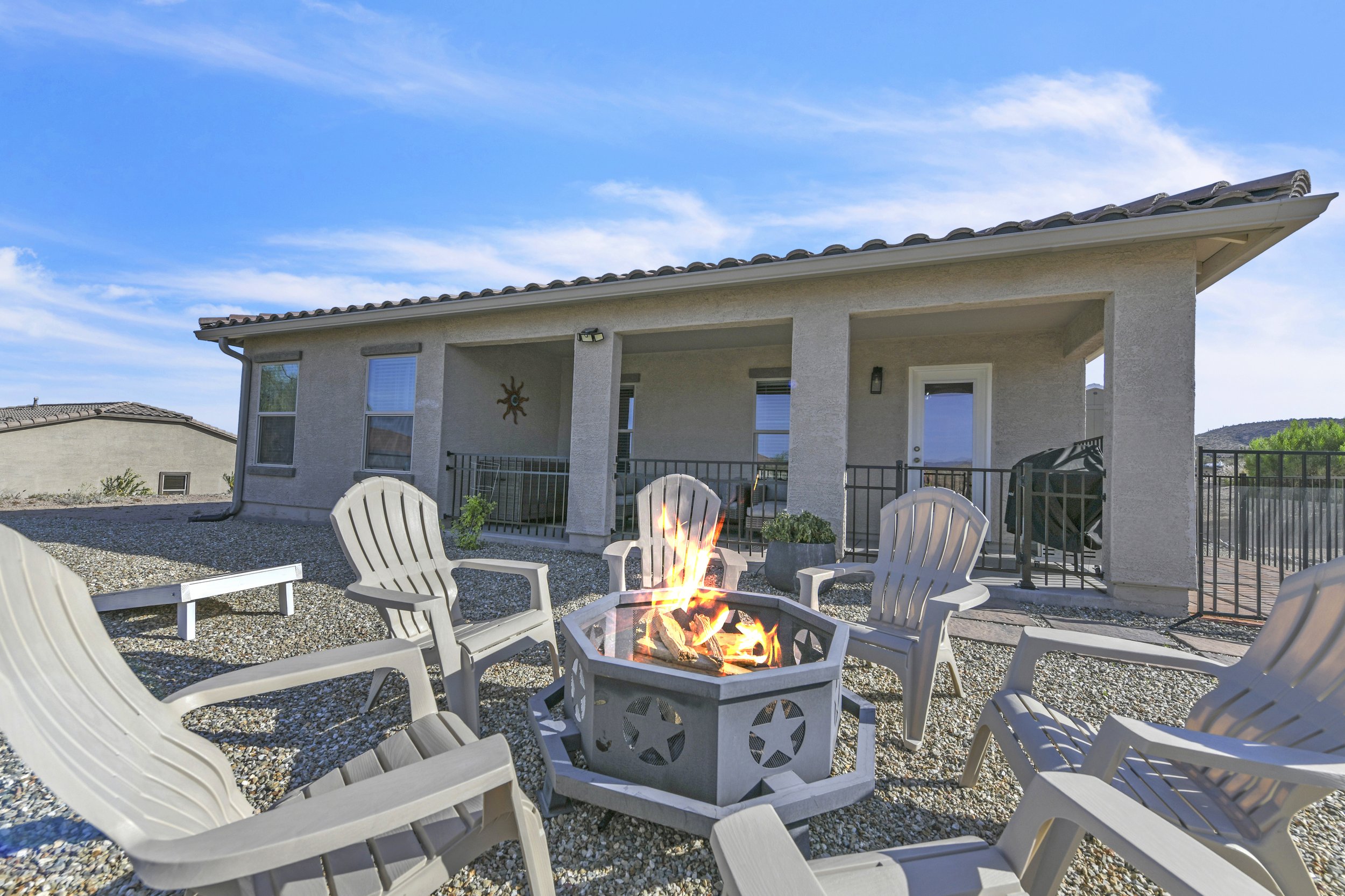 Backyard patio with a fire pit surrounded by white plastic chairs, a small house with a covered porch, and mountains in the distance under a blue sky.