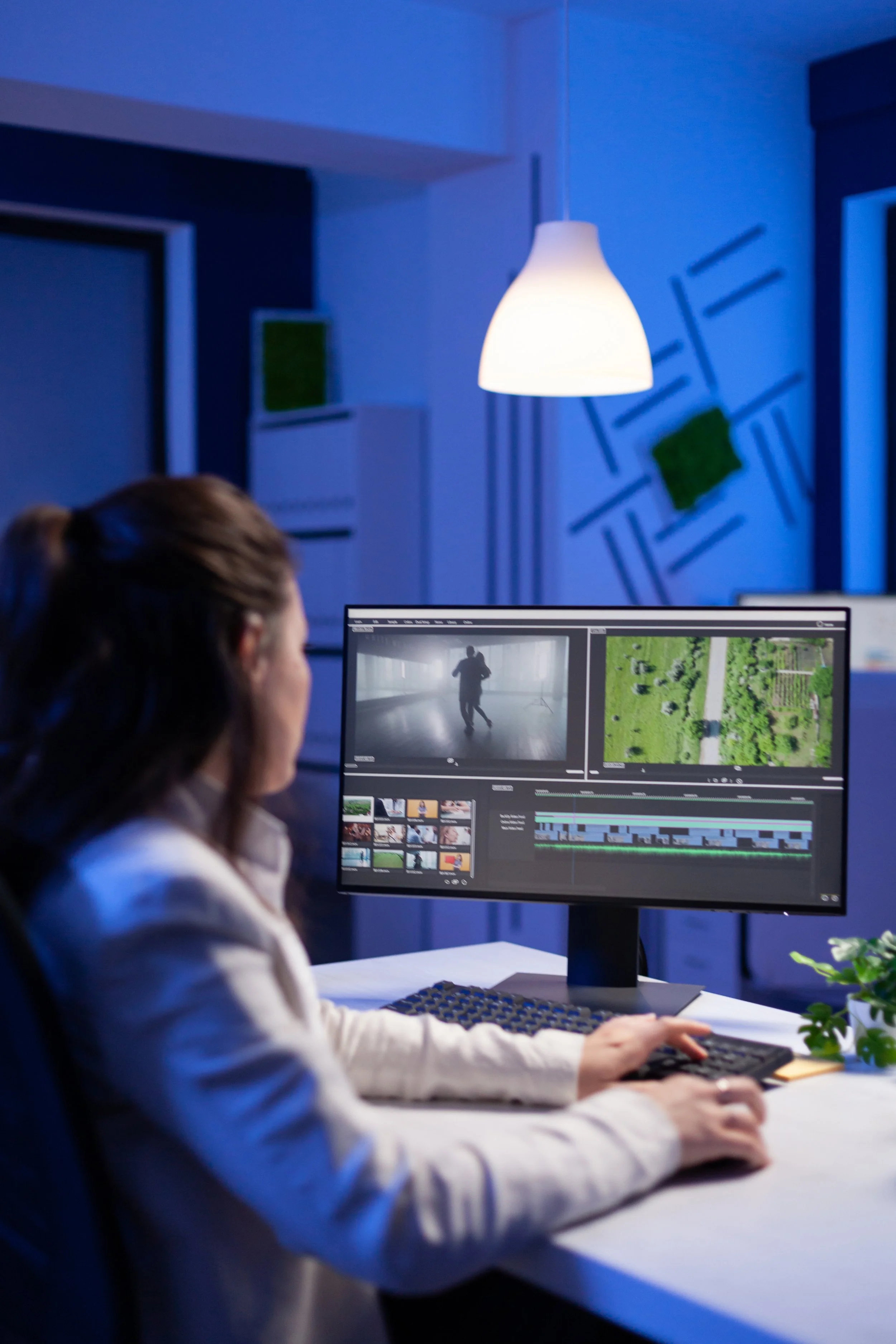 A person editing a video on a computer in a modern office with blue lighting. The monitor displays video editing software with a silhouette of a person running and aerial shots of a green landscape.