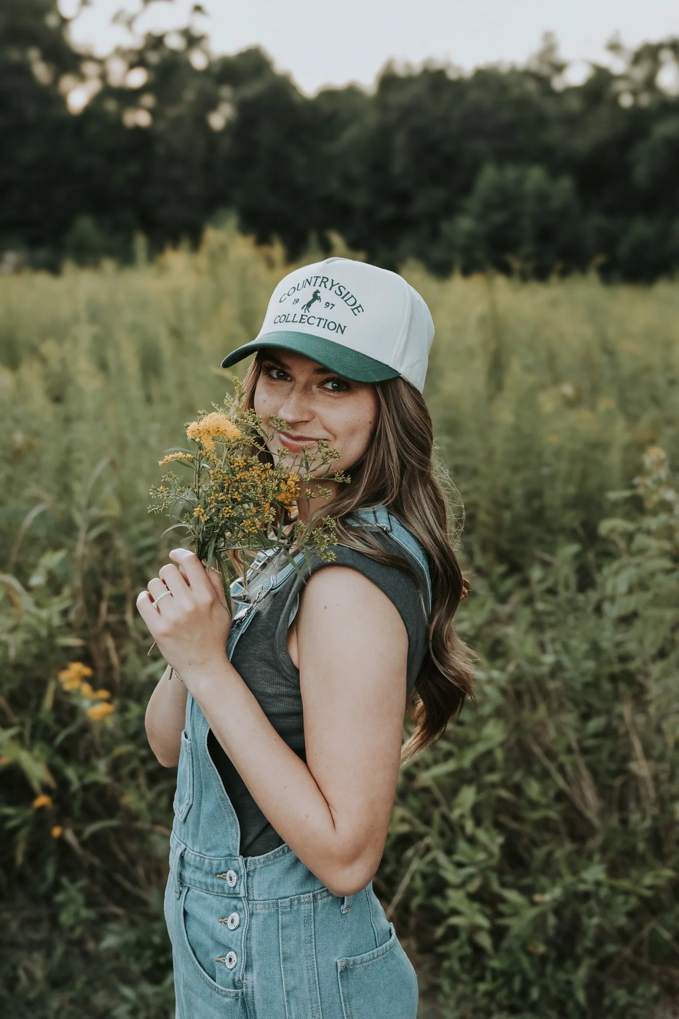 Sedona elopement photographer in overalls standing in a lush green backdrop for personal introduction.