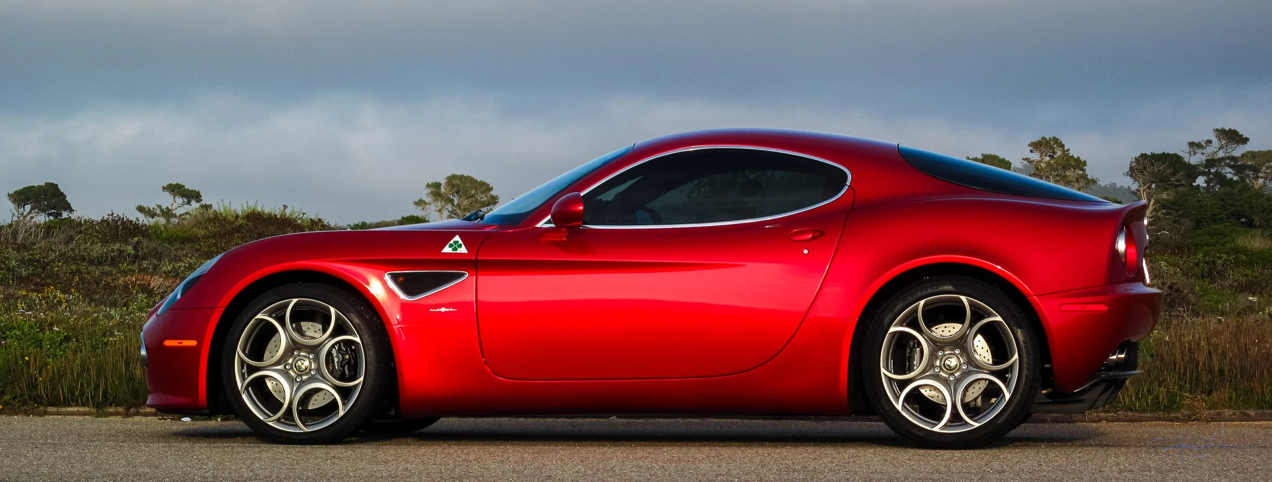 Red Alfa Romeo 8C sports car parked on a road with greenery and trees in the background.