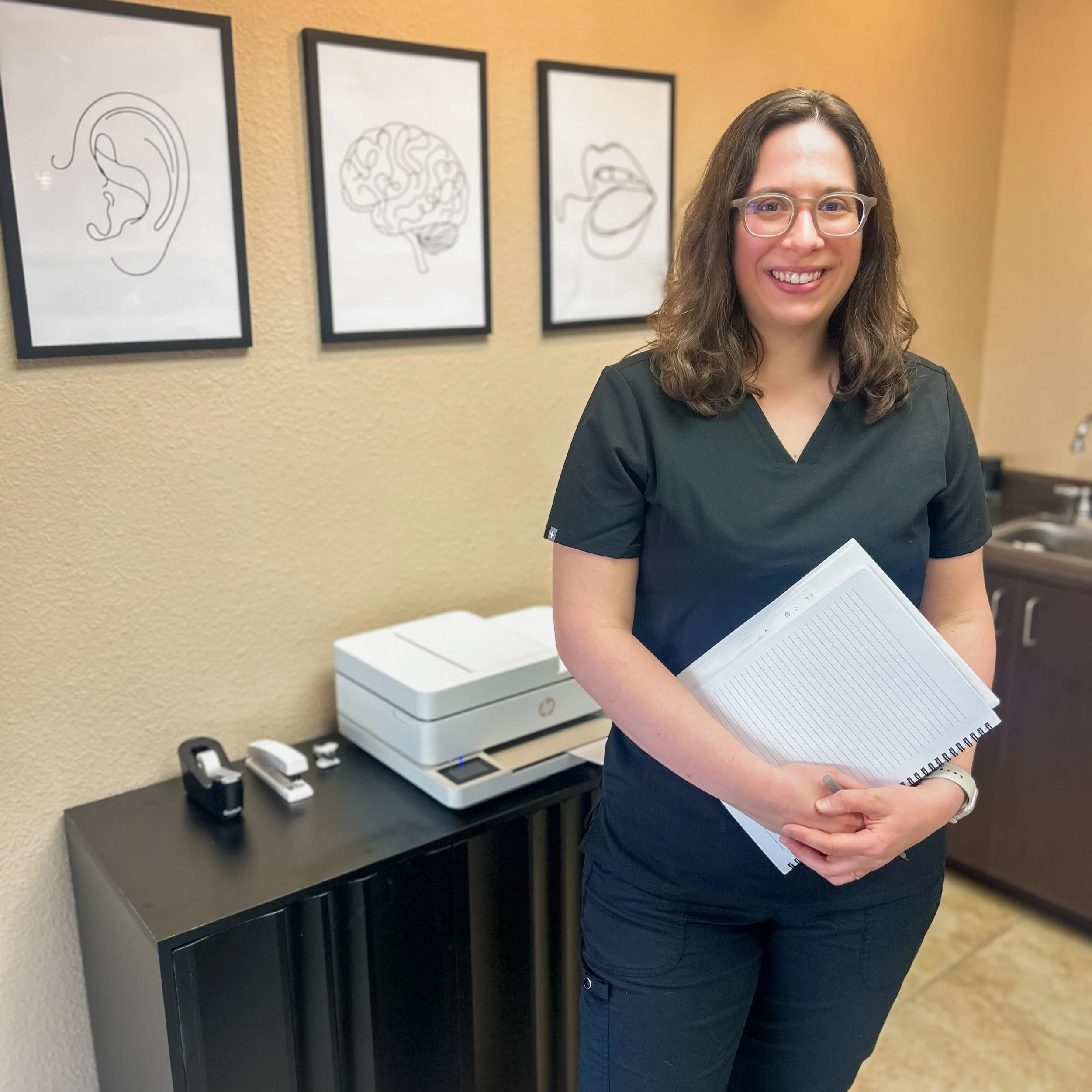 A woman with glasses and curly brown hair holding a notepad and pen, standing in an office or clinic with framed artwork of an ear, brain, and lips on the wall behind her.