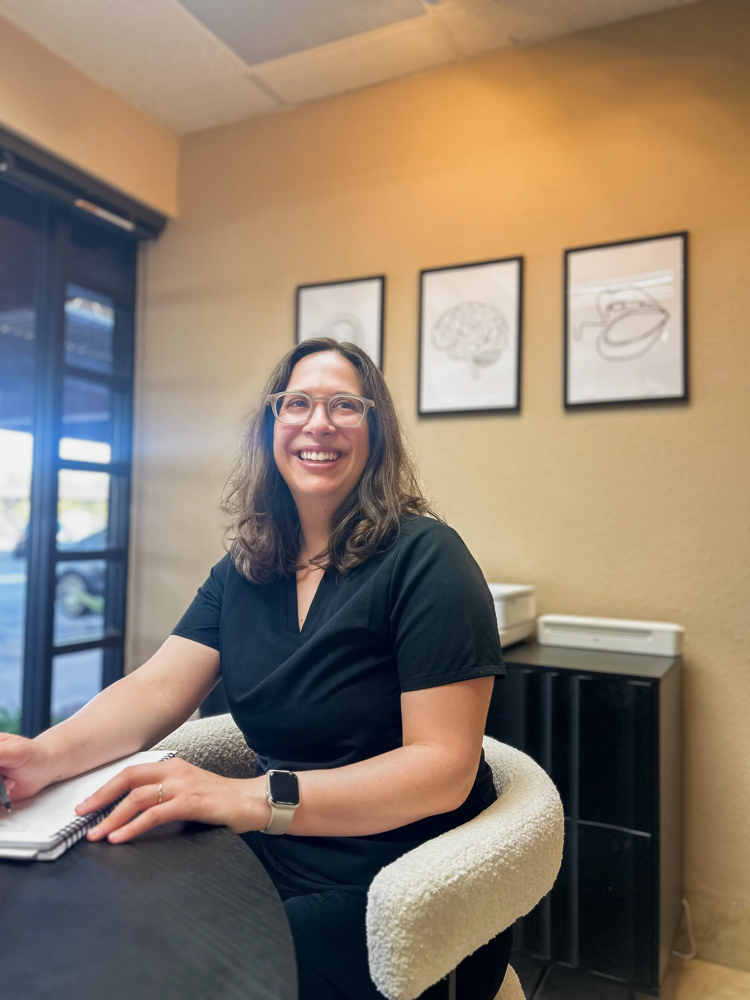 A woman wearing glasses and a smartwatch, smiling while sitting at a table, with a notebook in front of her in a room with framed art on the wall.