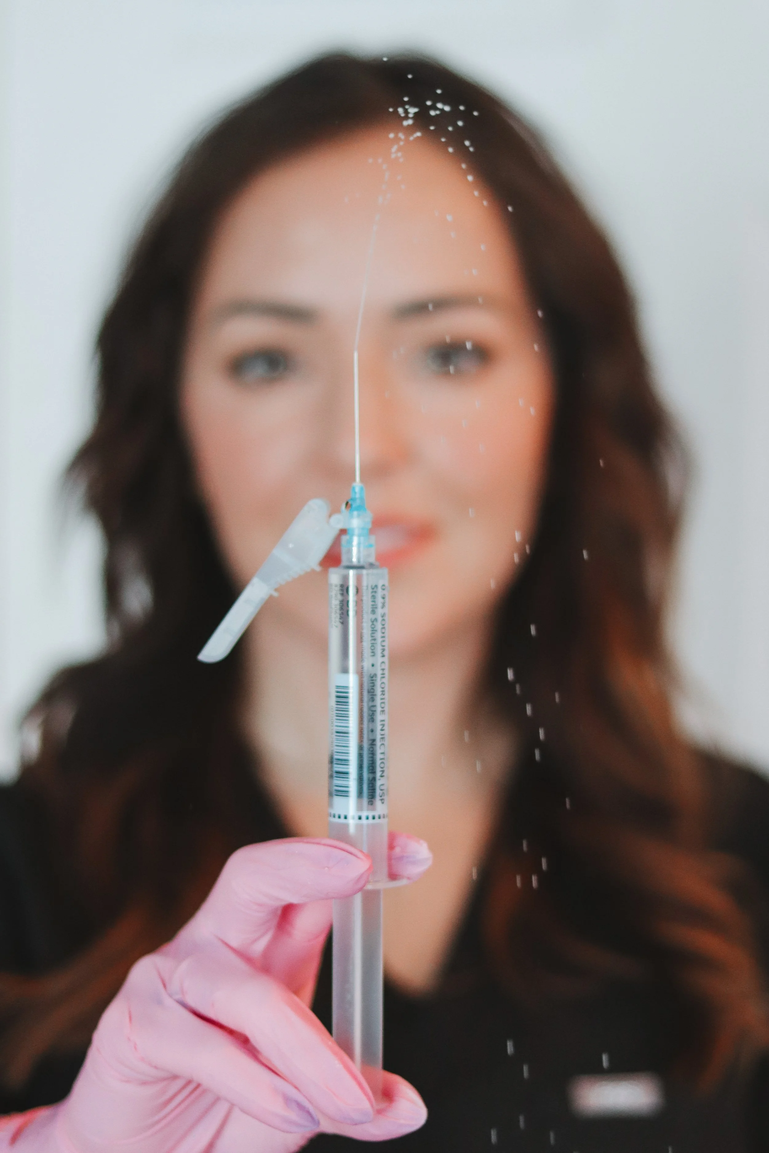 A healthcare worker with pink gloves holds a syringe aimed at the camera, with a woman blurred in the background, possibly about to receive a vaccine.