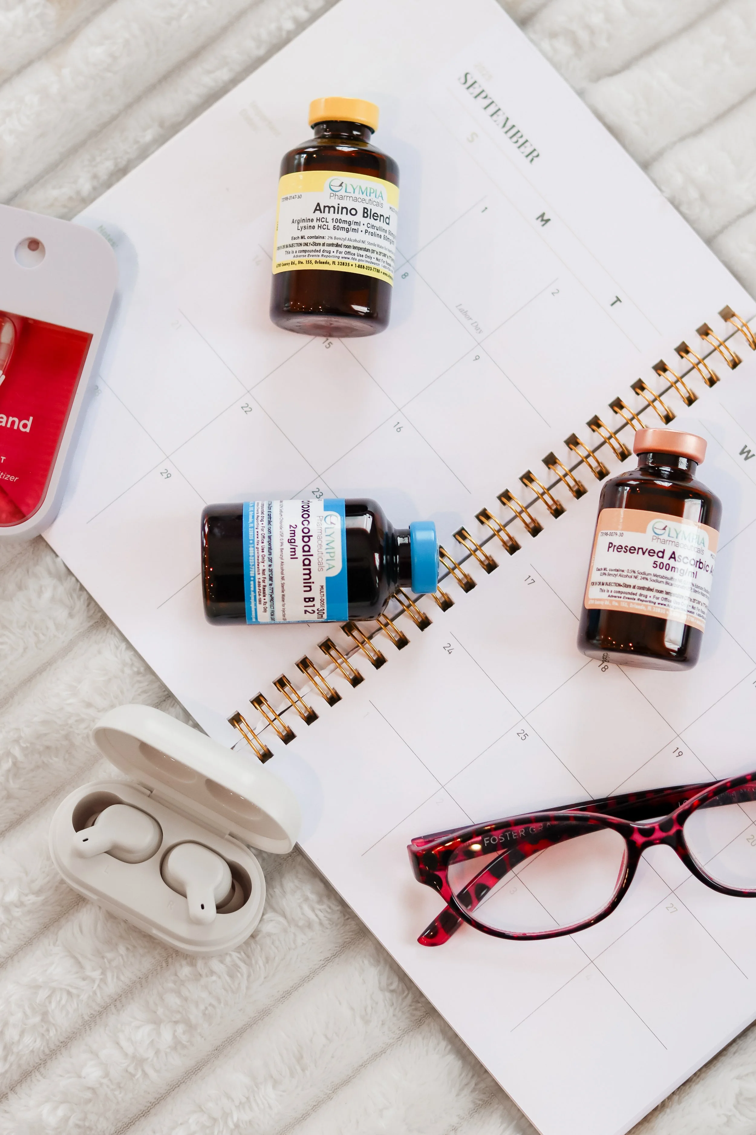 A planner or calendar with September open, three bottles of supplements or medications, a pair of red and black glasses, wireless earbuds in a white case, and a red item in the top left corner on a soft white surface.