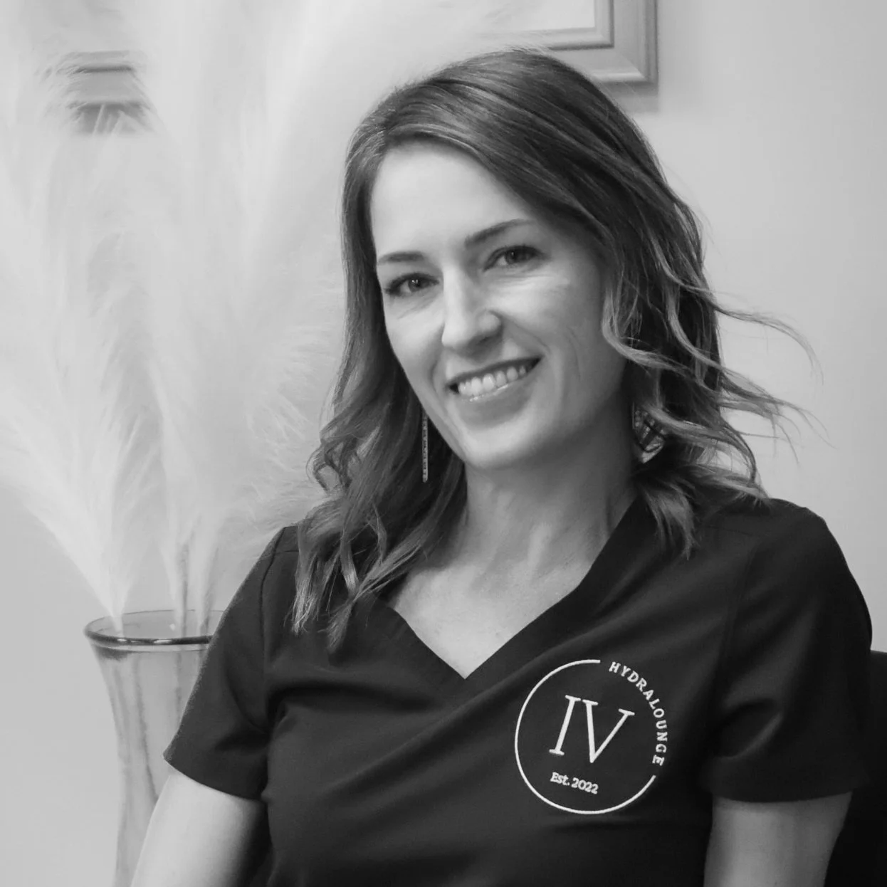 A woman with medium-length wavy hair smiling at the camera, wearing a dark shirt with a logo that reads "Hydralounge" and "Est. 2022." in front of a plain background with a potted plant.
