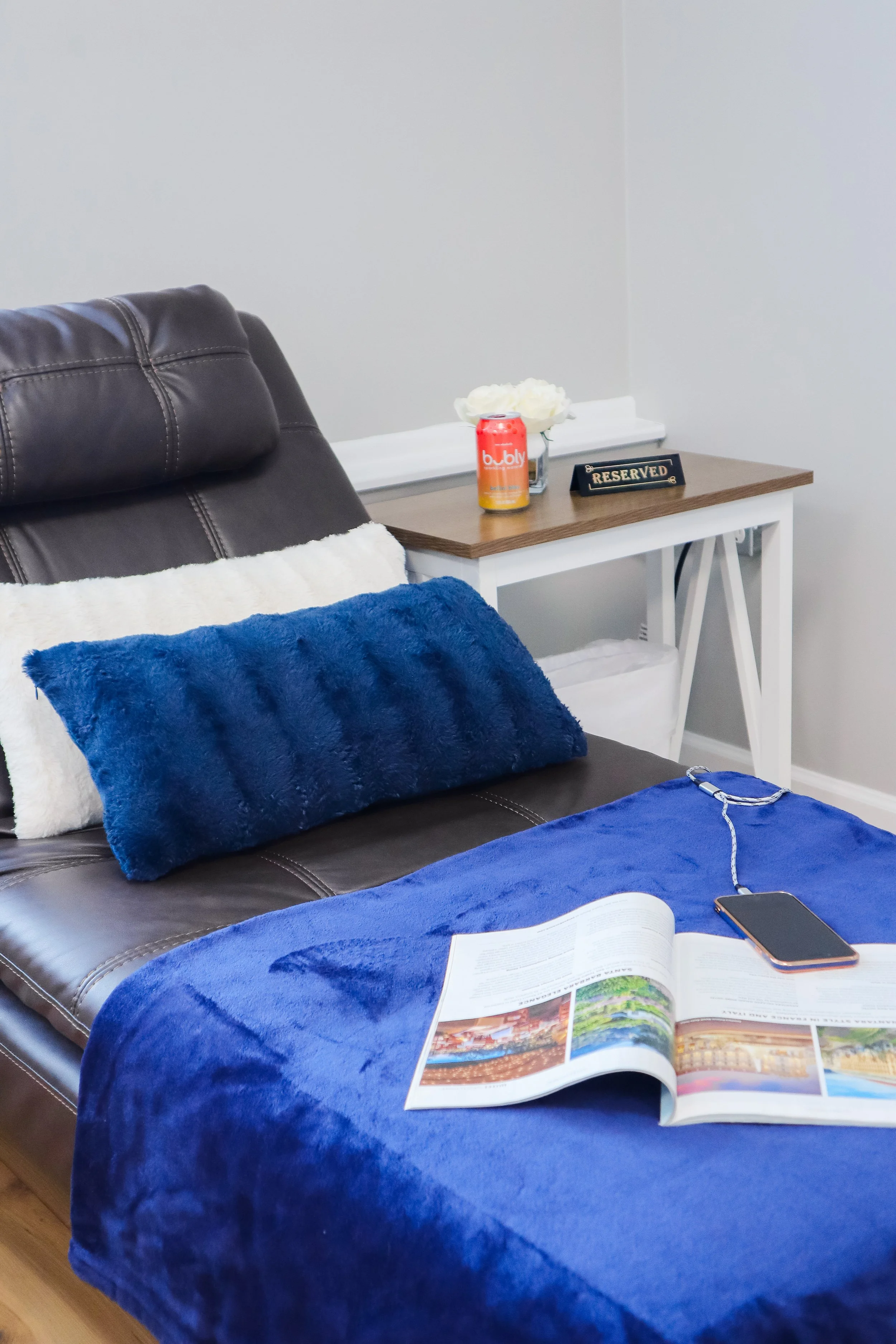 A living room setup with a brown leather couch covered with a fluffy white pillow and a blue fuzzy pillow. A blue blanket is draped over the couch. A magazine is open on the blanket, and a smartphone with a charger is placed beside it. Behind the cou