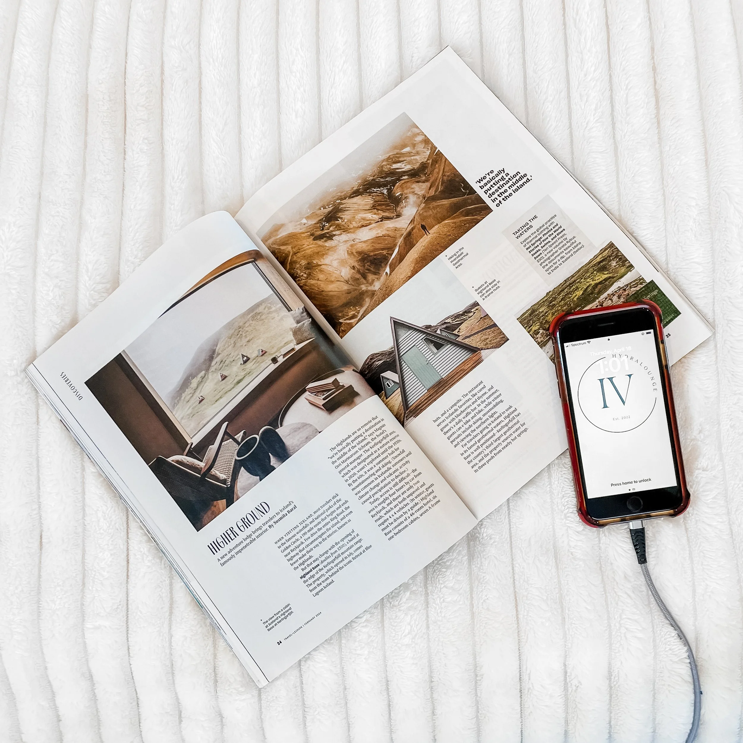 Open magazine with landscape photos and articles, placed on a white textured cloth, with an iPhone connected to a charging cable displaying a screen with the text 'TOP IV'.