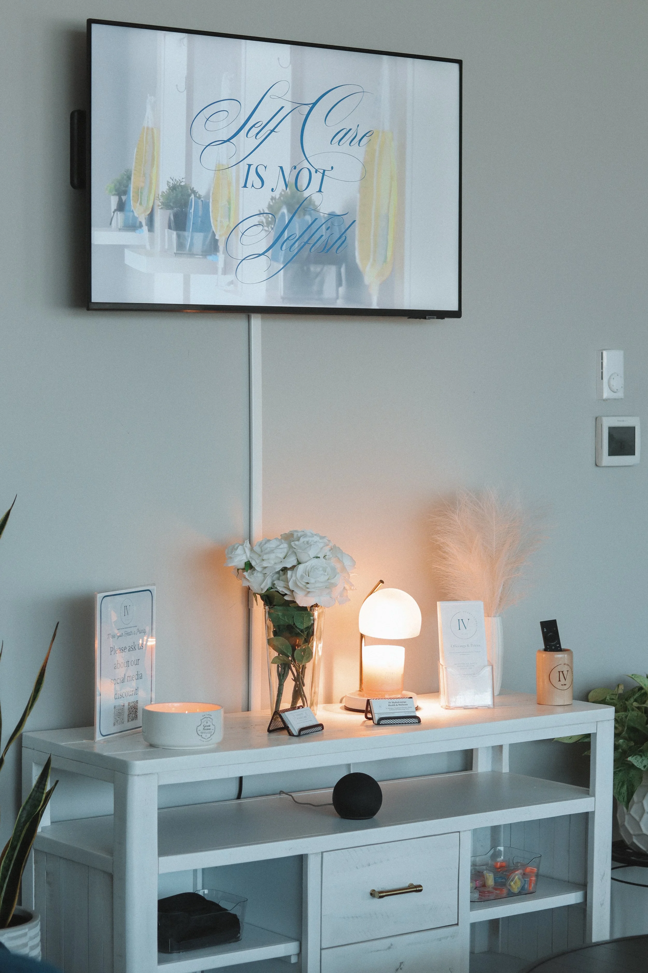 A white table with a flower vase, a white lamp, and informational cards, in front of a wall with a mounted TV screen displaying the quote "Self Care is Not Selfish".