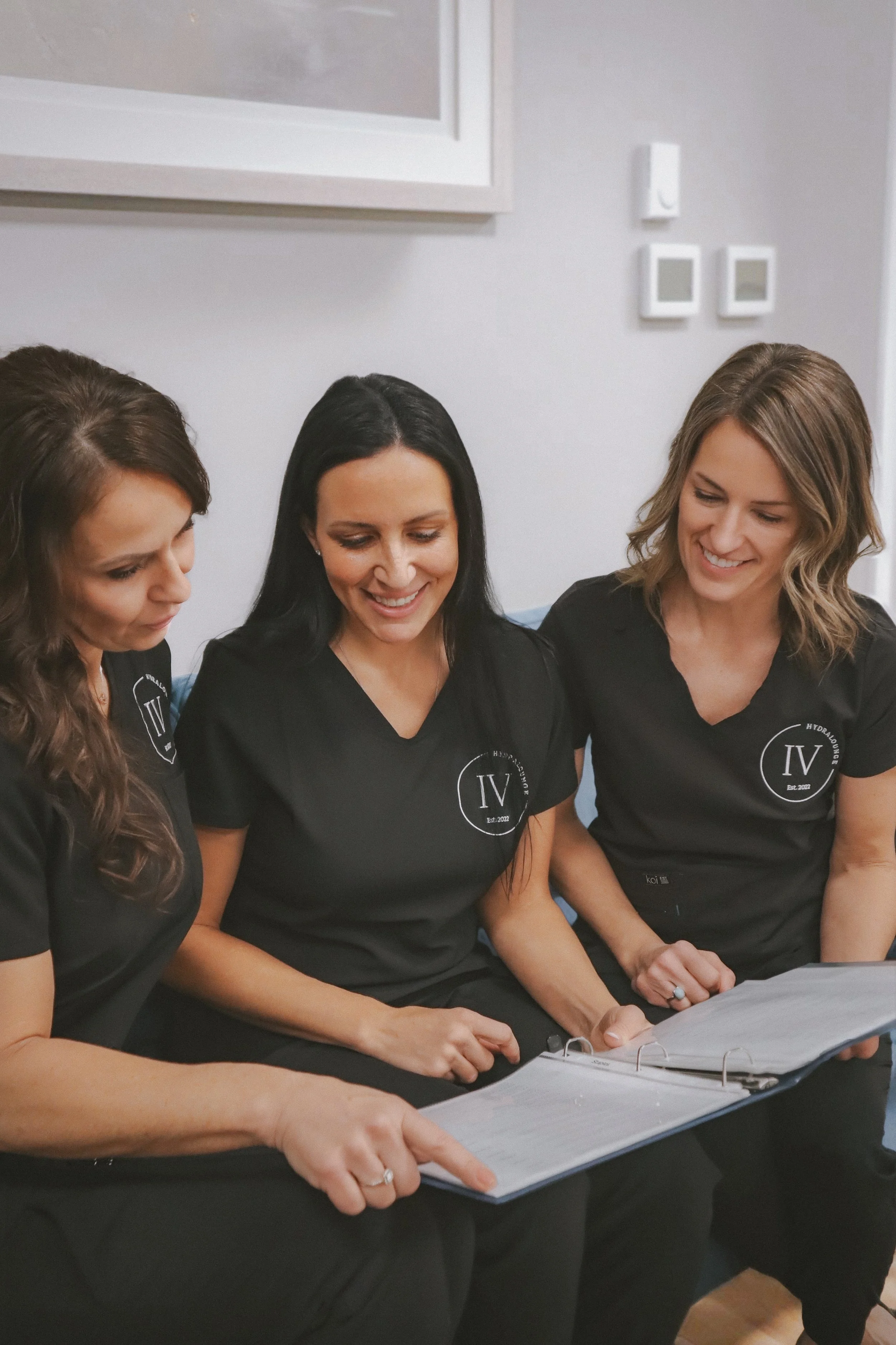 Three women in black uniforms sitting on a bench with a binder, engaged in conversation and smiling