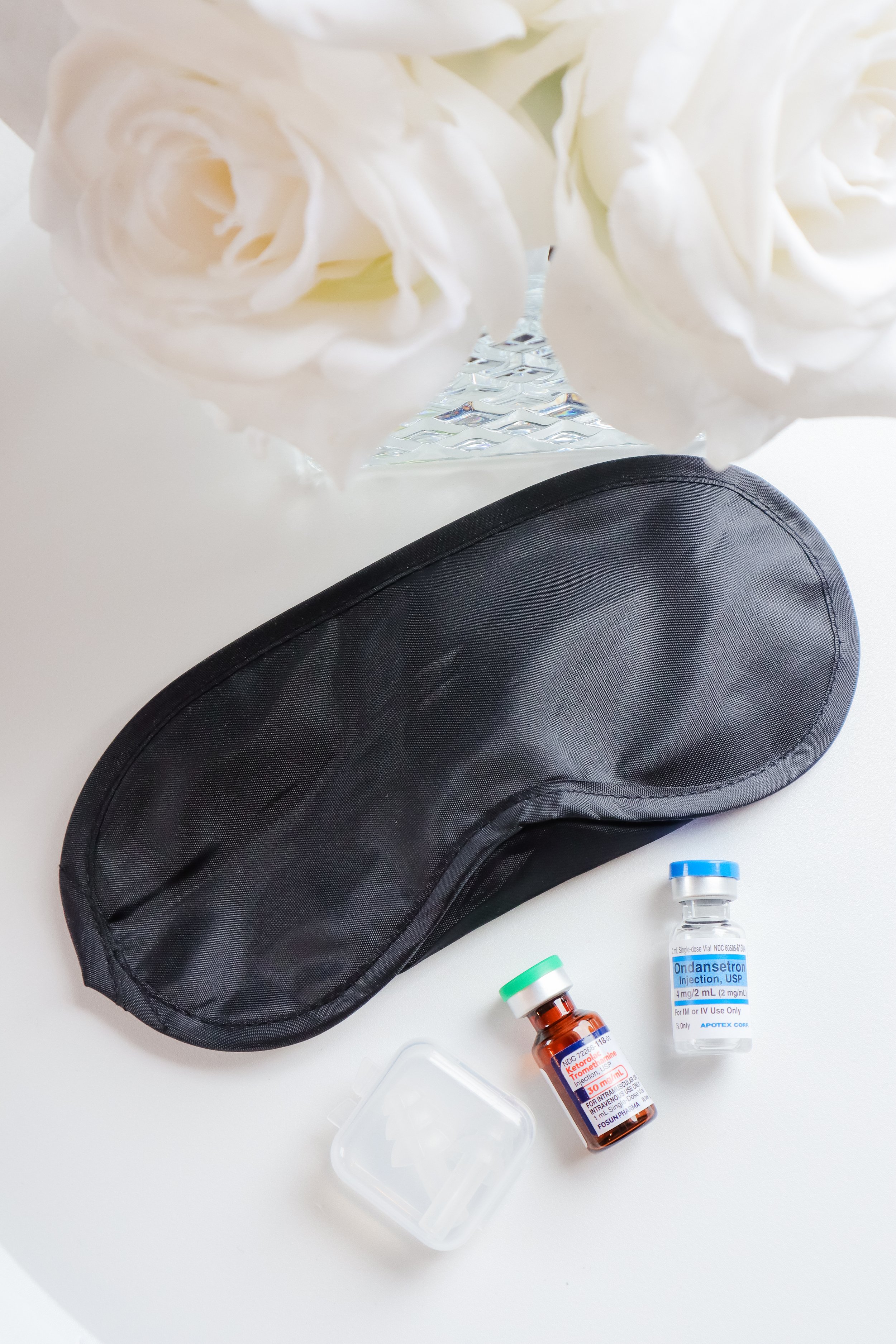 A black sleep mask, two insulin vials, and a small plastic container on a white surface, with white roses in a glass vase in the background.