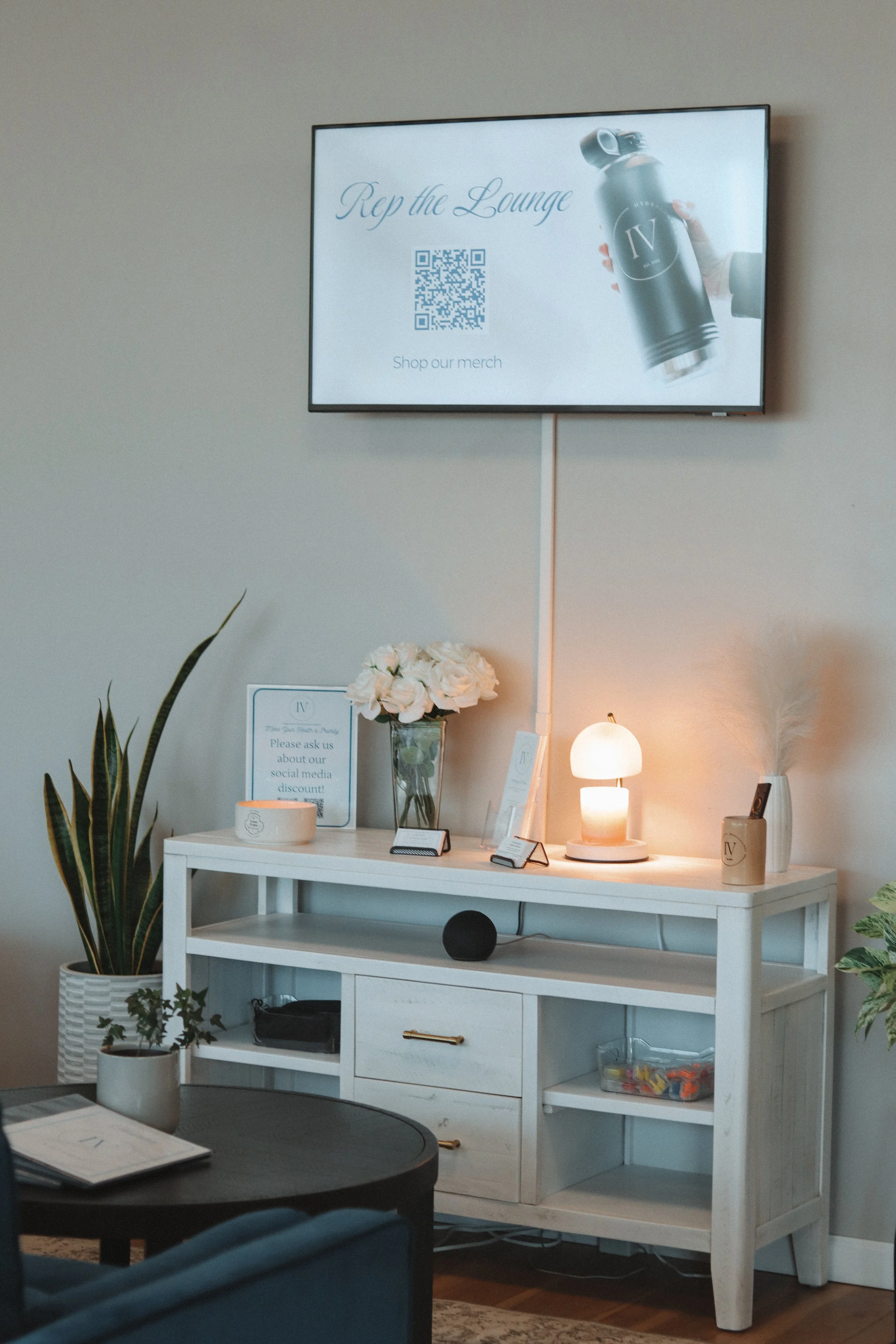 A lobby or lounge area featuring a white console table with a potted plant, a sign, a vase with white flowers, a lamp, and a small white vase. There are small display cards and a speaker on the table. Mounted on the wall above is a TV screen promotin