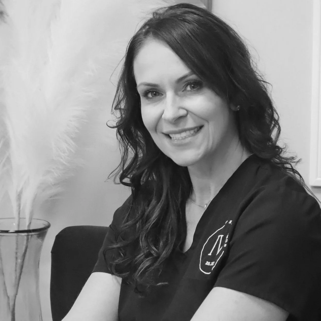 A smiling woman with dark curly hair wearing a dark shirt, sitting with her arms crossed, next to a tall vase with white feathers.