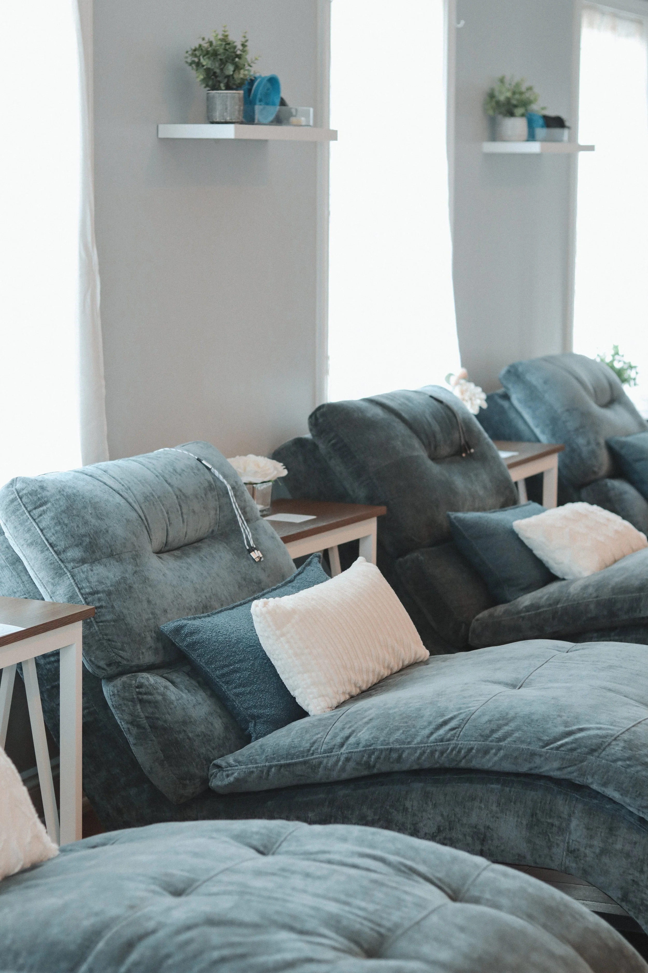 Multiple gray plush lounge chairs with pillows arranged in a row in a bright room with large windows and white curtains. Small side tables with decorative items and potted plants are placed near the chairs.