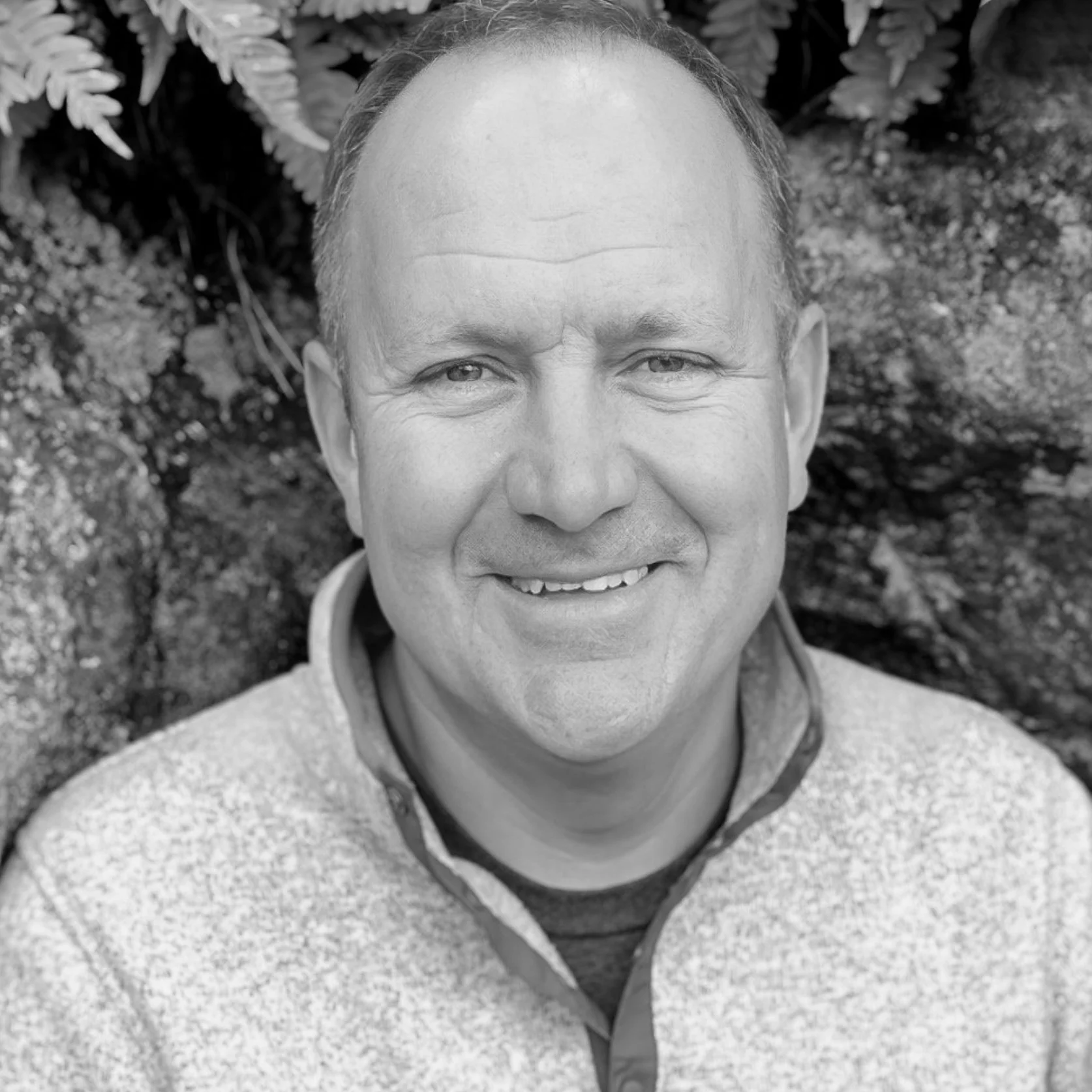 A black-and-white photograph of a smiling middle-aged man with short hair, outdoors with a rocky background and some foliage.