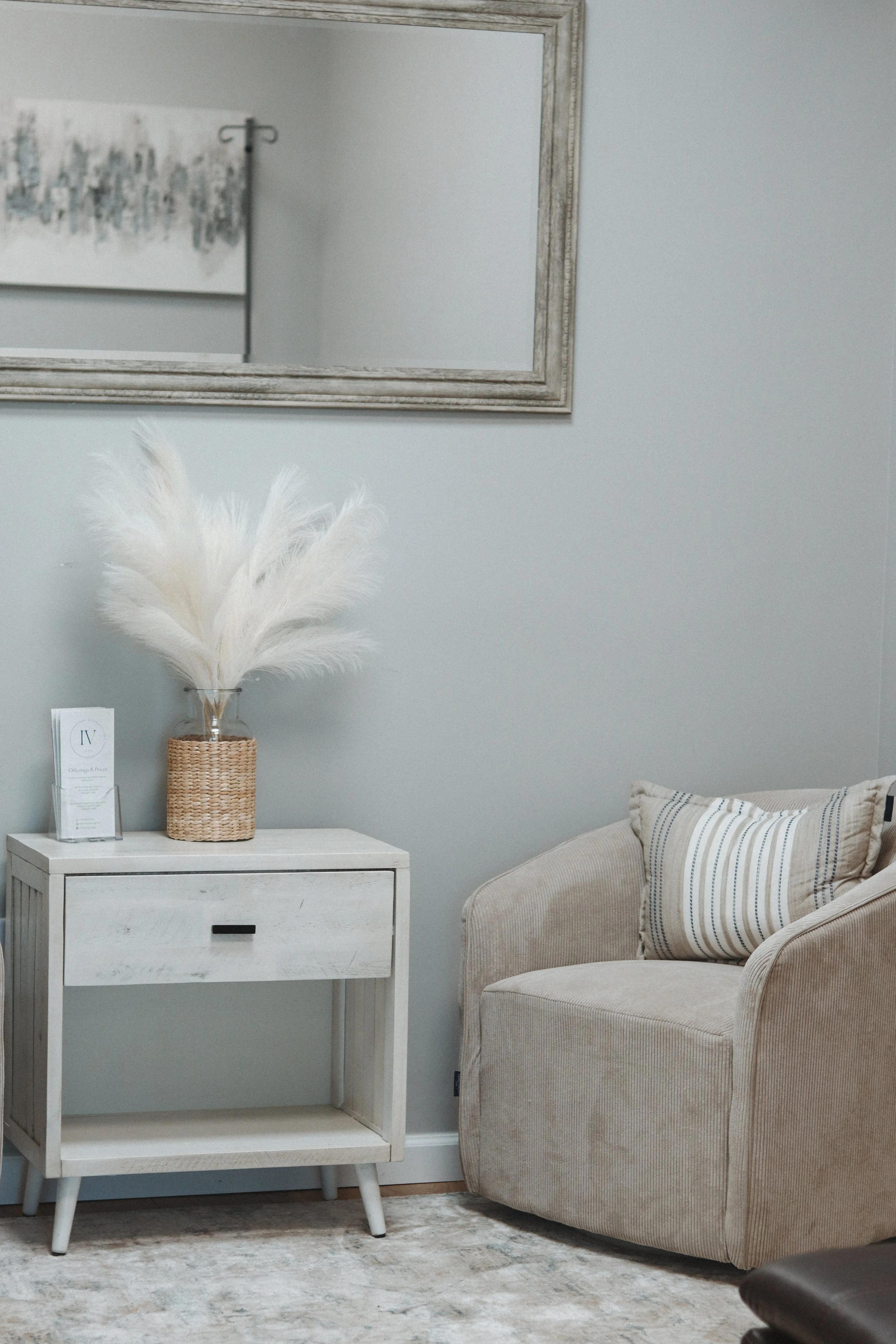 A cozy living room corner featuring a beige armchair with a decorative pillow, a white wooden side table with a drawer, a wicker vase with white pampas grass, and a large mirror on the light blue wall.