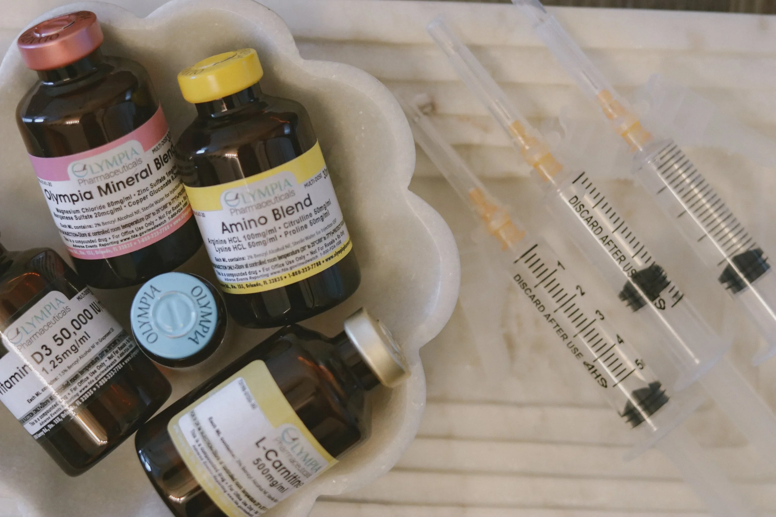 Medical supplies including vials labeled Olympia Pharmaceuticals for mineral, amino, and vitamin blends, and syringes in plastic packaging.