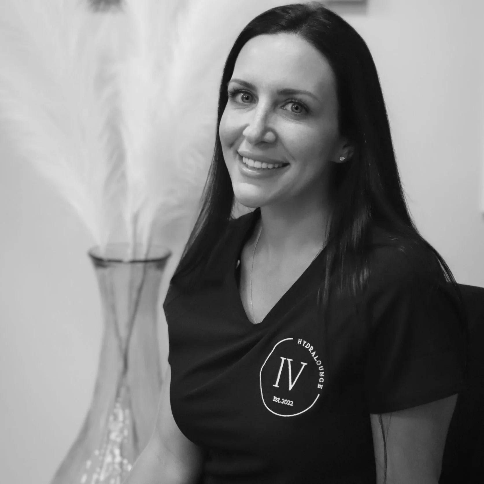 A woman with long dark hair smiling at the camera, wearing a black shirt with the logo 'Hydralounge IV Est. 2022' on it, sitting indoors with a decorative vase with tall feathers in the background.