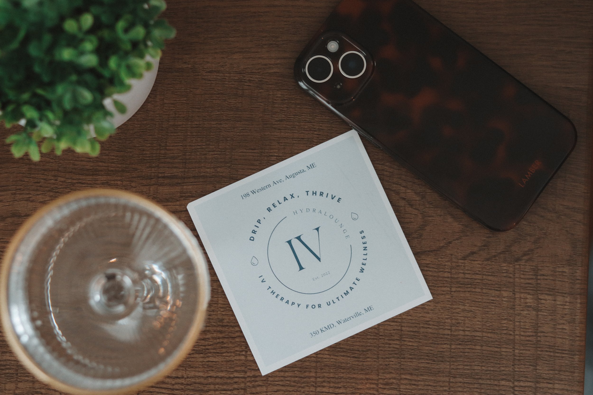 A top view of a wooden table with a green potted plant, a glass of water, a printed flyer, and a smartphone.