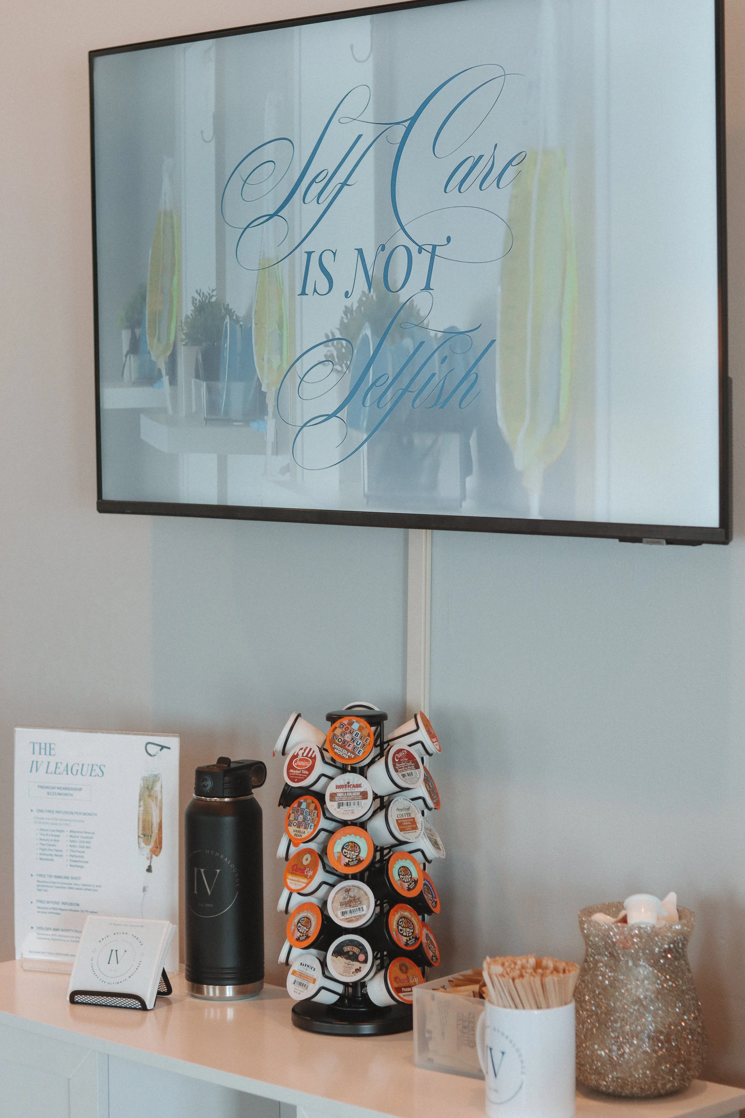 A display counter with a large screen behind it showing the quote "Self Care Is Not Selfish" in elegant cursive and block fonts. In front of the counter, there are several coffee pods on a carousel, a black water bottle with "IV" on it, a stand holdi
