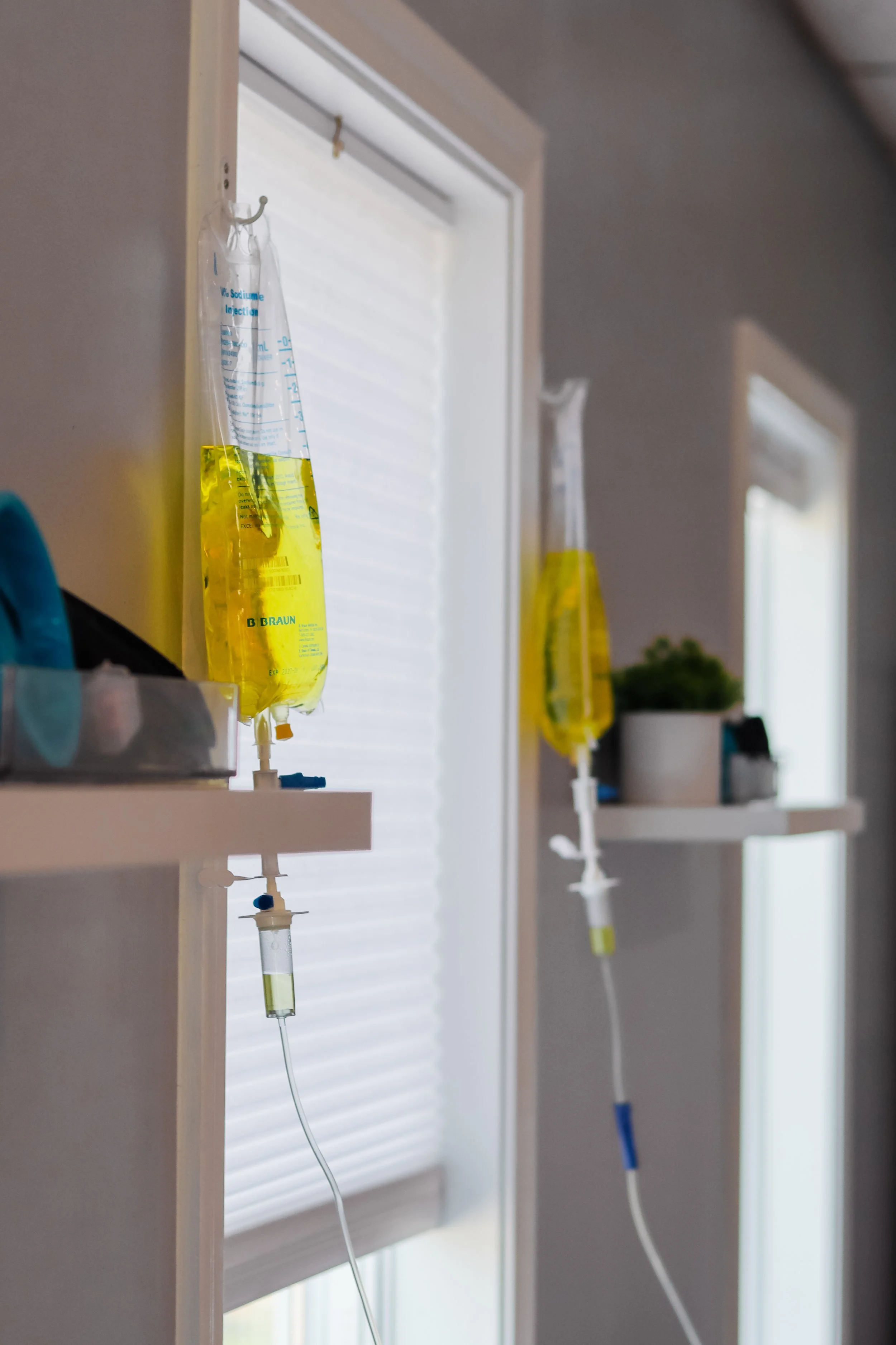 Two IV medication bags filled with yellow liquid hanging on hooks in a hospital room near a window.