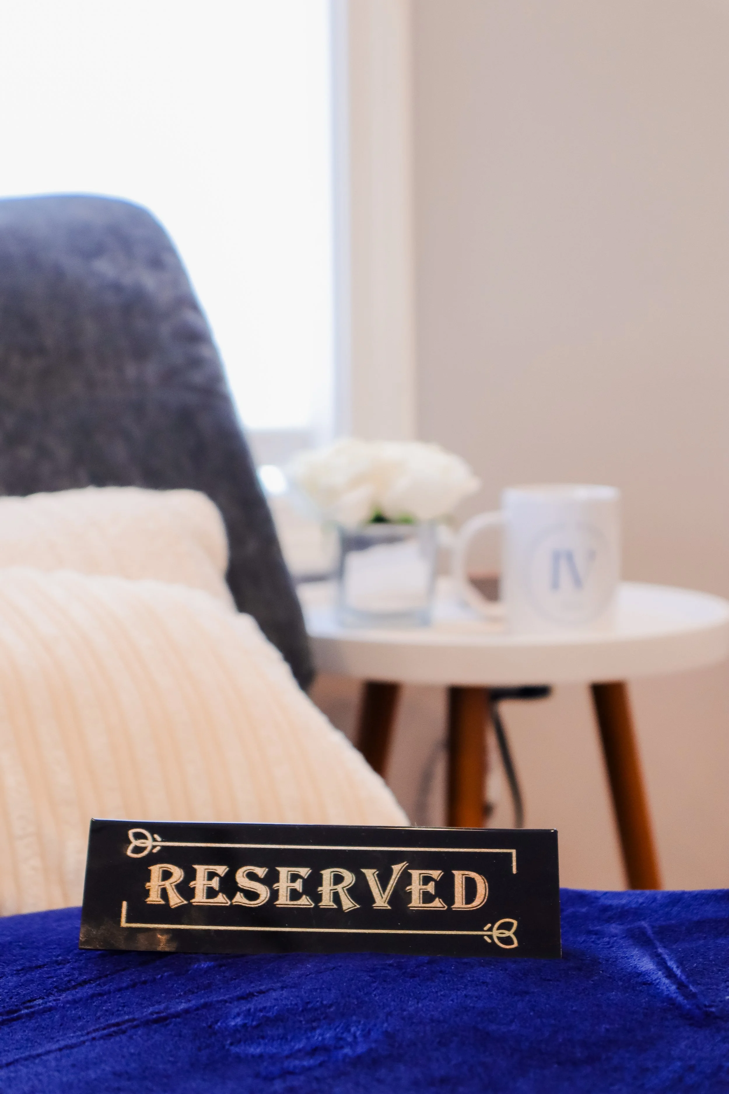 A reserved sign placed on a blue fabric surface in the foreground. In the background, there is a white side table with a white mug, a flower vase with white flowers, a pillow, and a window.