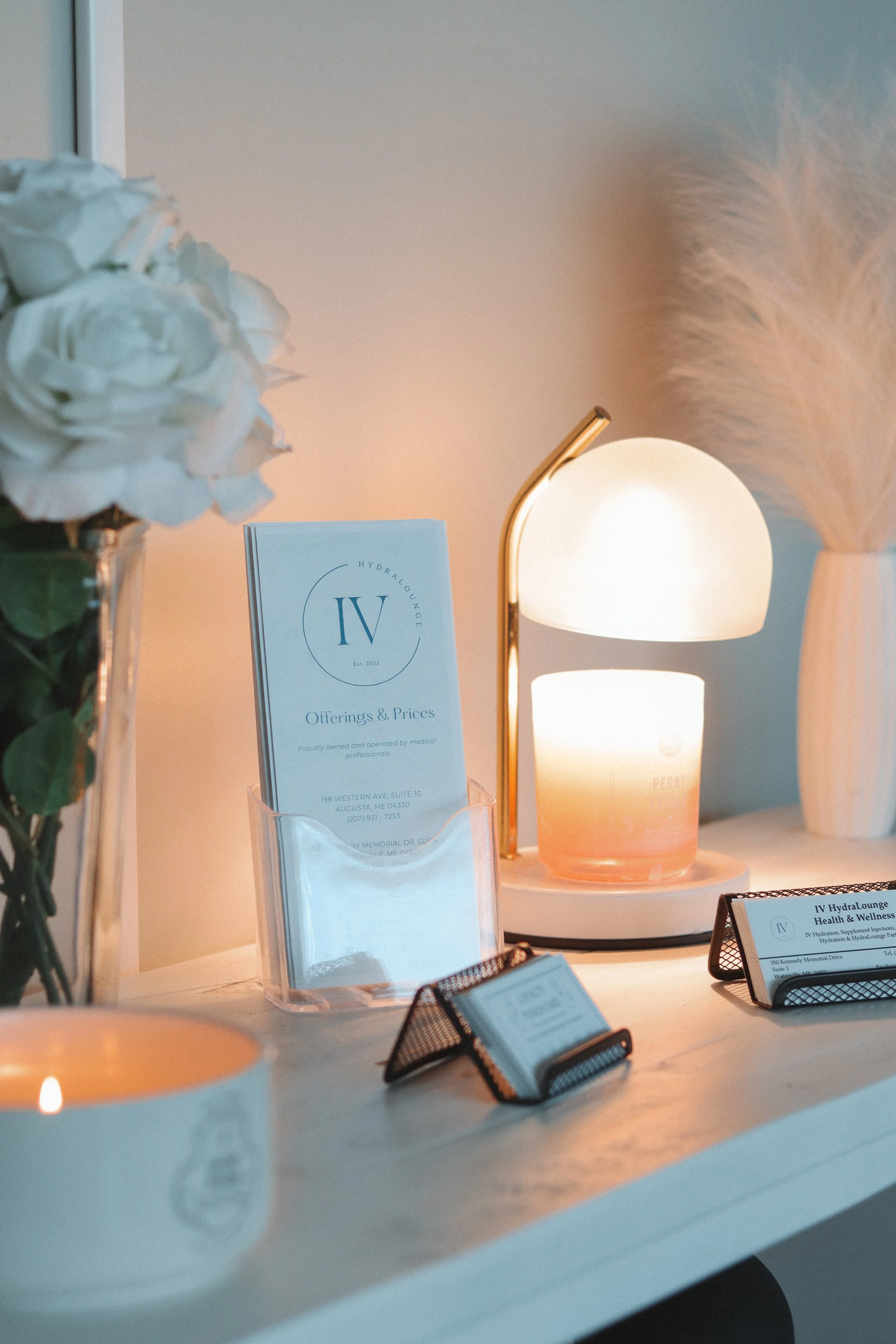 A white table with a small lampshade, a vase with pampas grass, a booklet titled 'Offerings & Prices', and small digital displays, in a softly lit room.