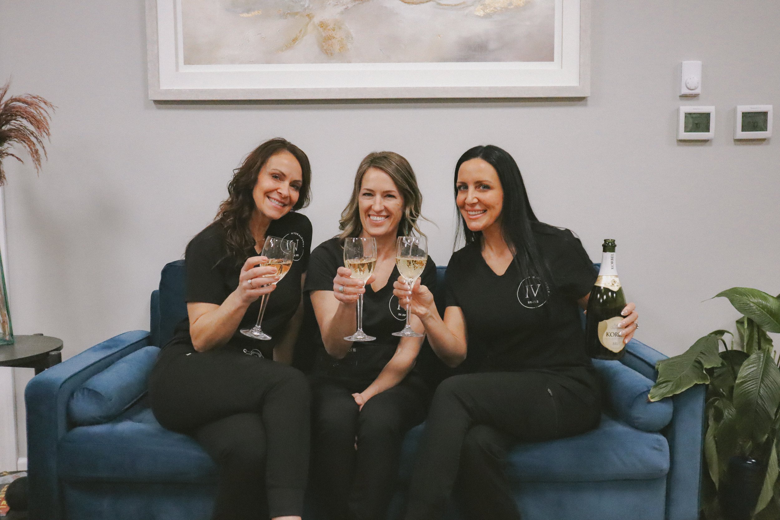 Three women sitting on a blue sofa, smiling, holding glasses of white wine, with one woman also holding a bottle of sparkling wine, in a well-decorated indoor setting.